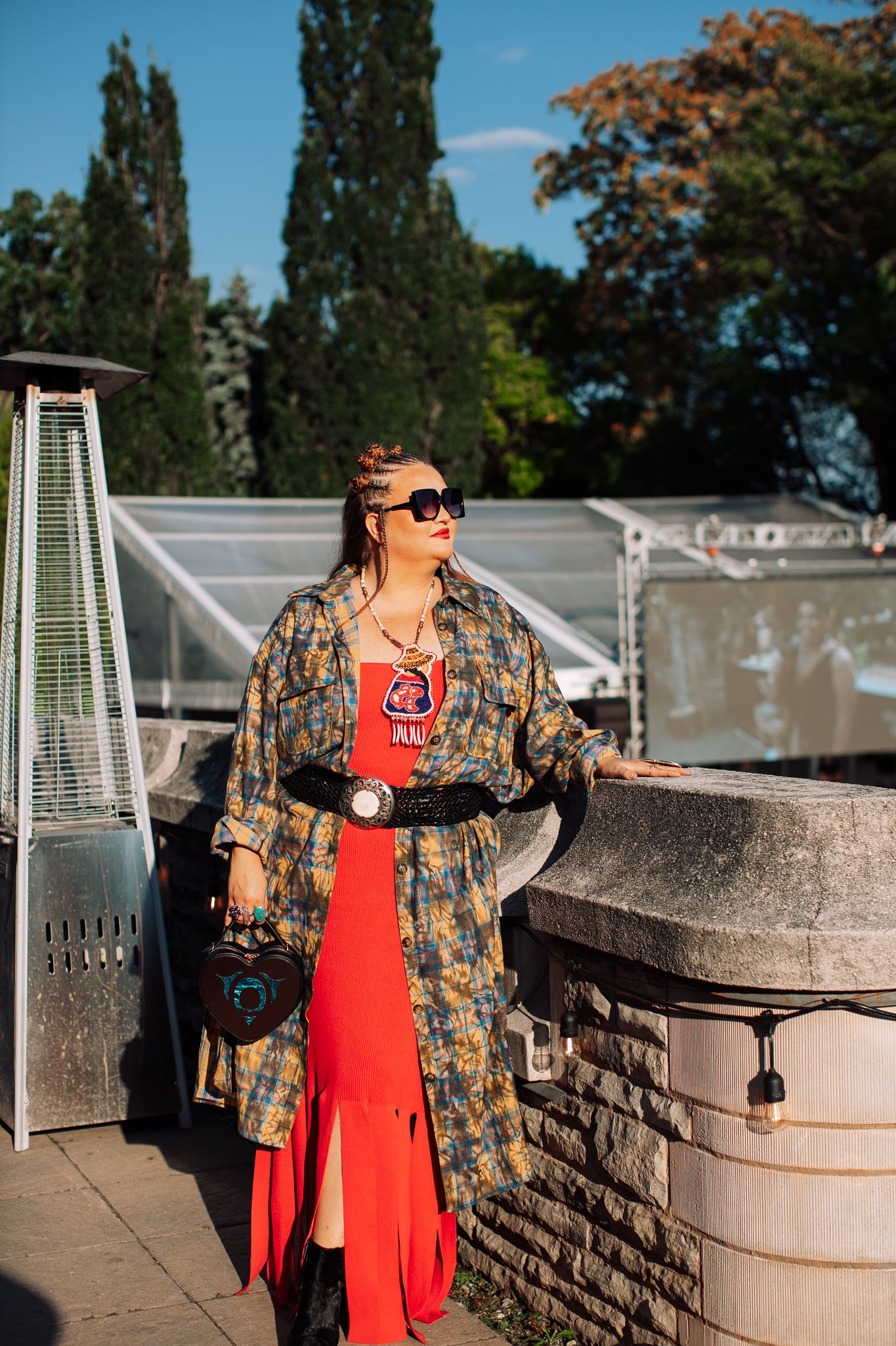 Woman in floral coat, red dress, sunglasses, and belt bag poses outdoors at a sunny event venue.