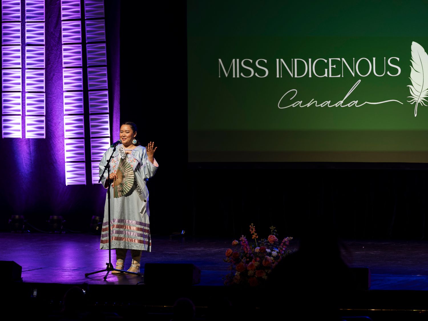 Performer speaks on stage at Miss Indigenous Canada pageant before a green feather logo backdrop.