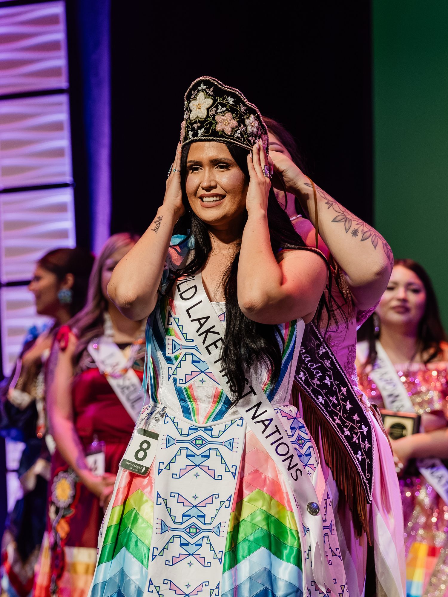 Miss Indigenous Canada contestant adjusts her crown on stage wearing a colorful regalia dress and sash.