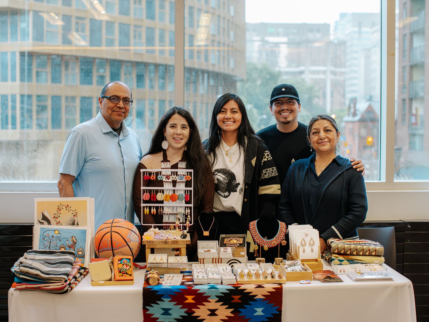 Five people smiling behind a vendor table displaying jewelry, crafts, and Indigenous goods at an indoor market event.