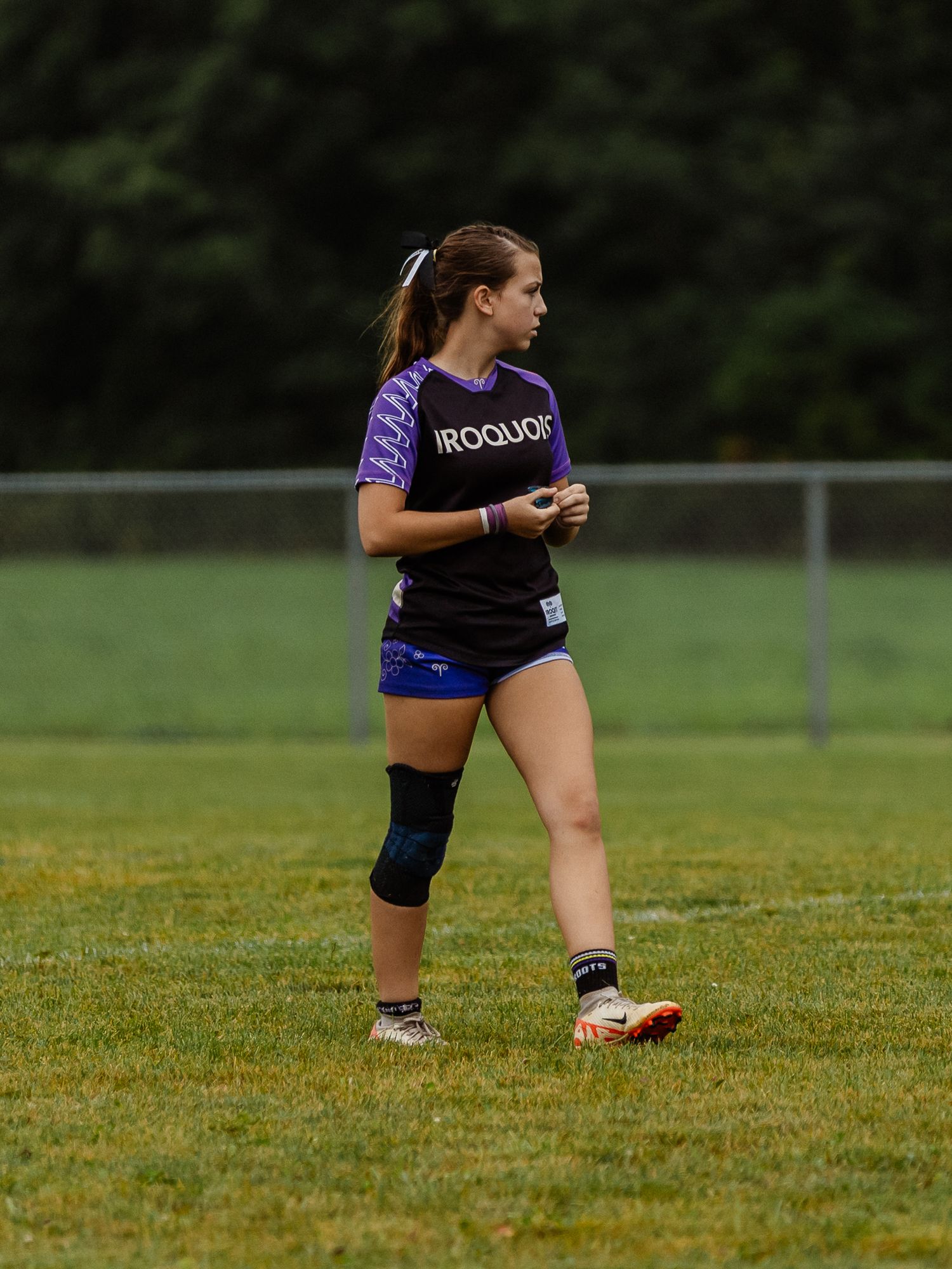 Female rugby player in Iroquois jersey with knee brace stands on green field during overcast game.