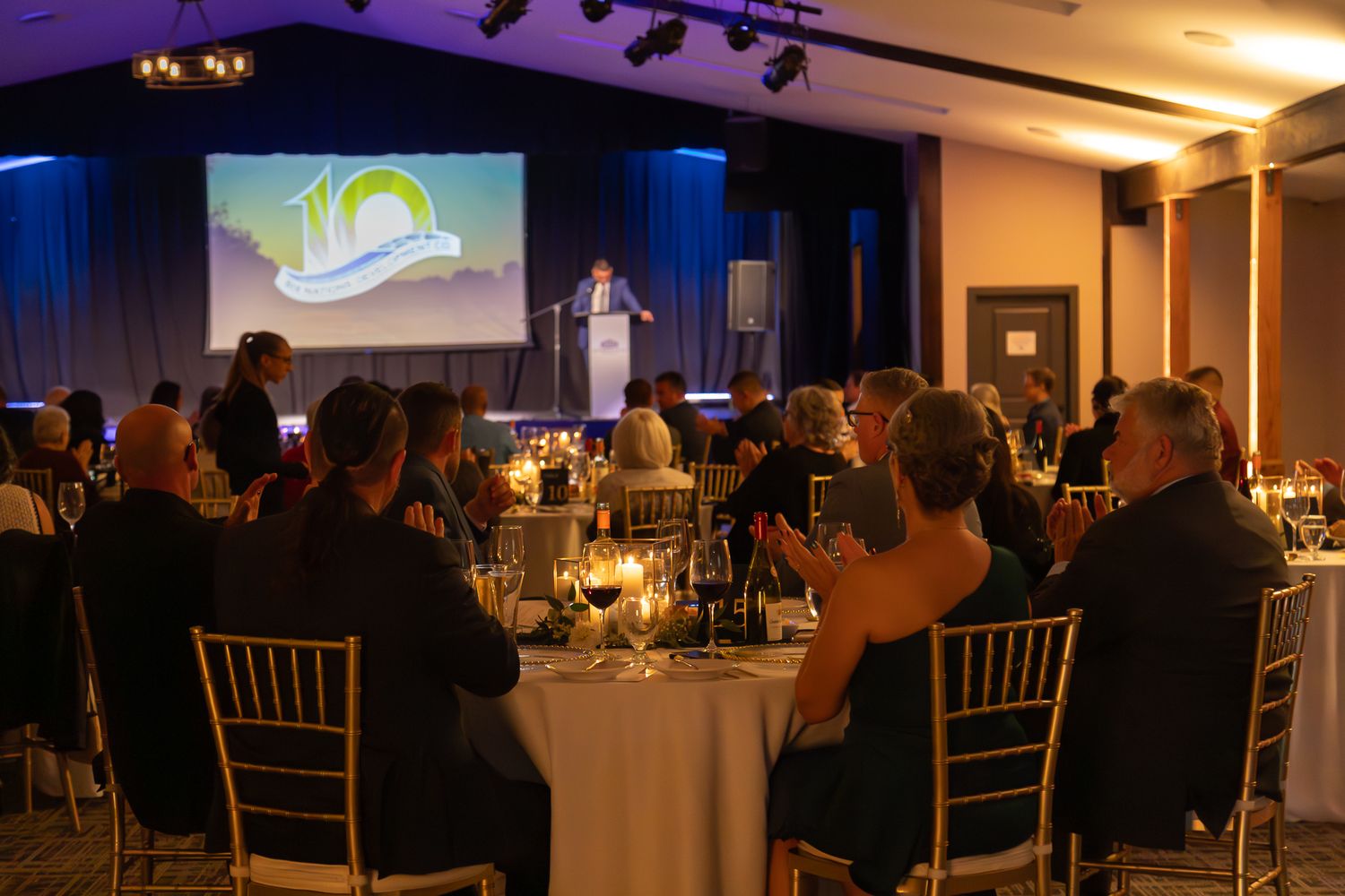Guests applaud at a formal gala dinner celebrating a 10th anniversary, with a speaker at the podium on a blue-lit stage.