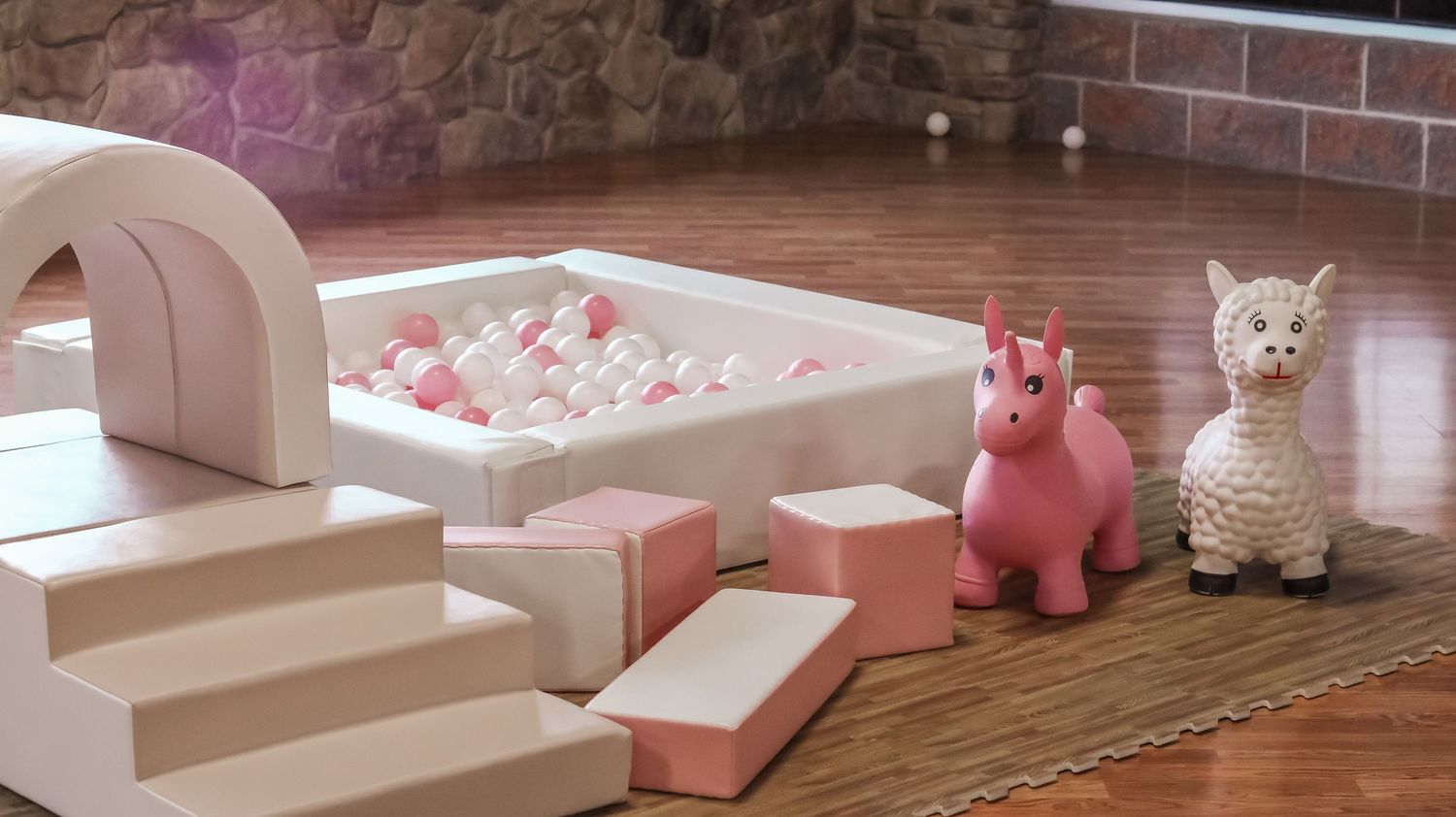 Pink and white foam play area with ball pit, building blocks, pink unicorn hopper, and llama toy on hardwood floor.