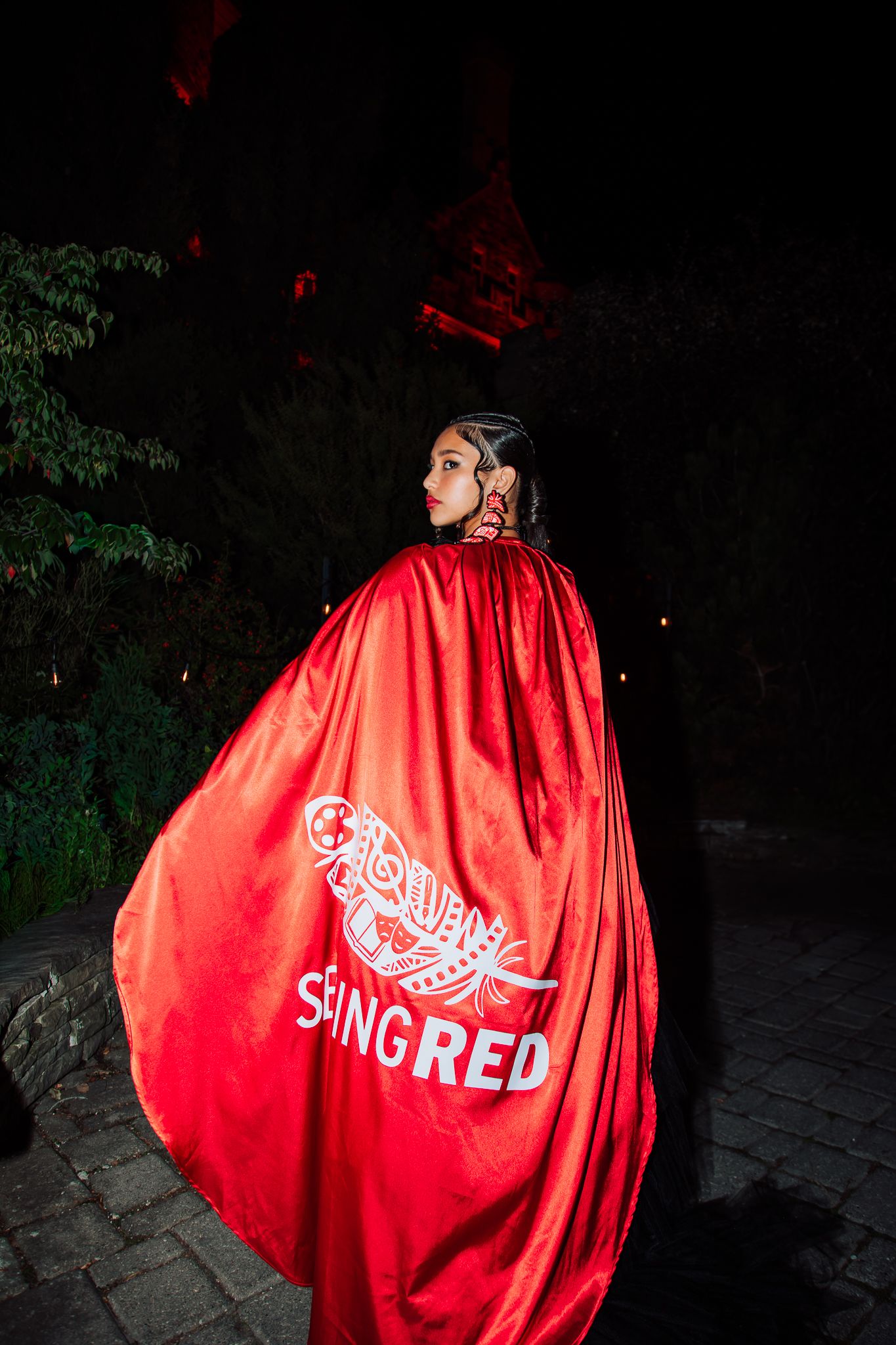 Person wearing a bright red cape with white logo design against dark outdoor background.