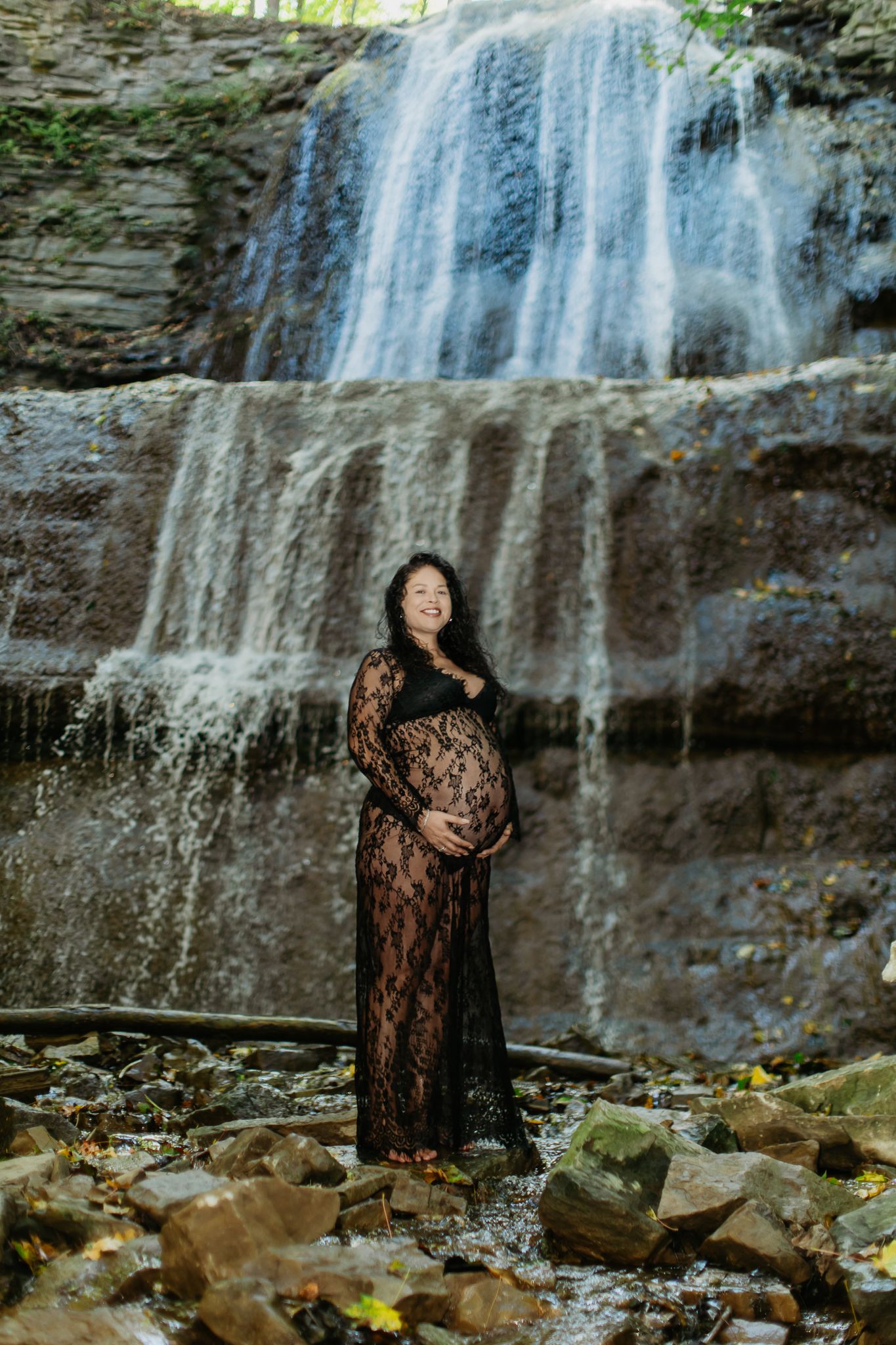 Artistic maternity portraits taken in front of a serene waterfall with natural rock formations.