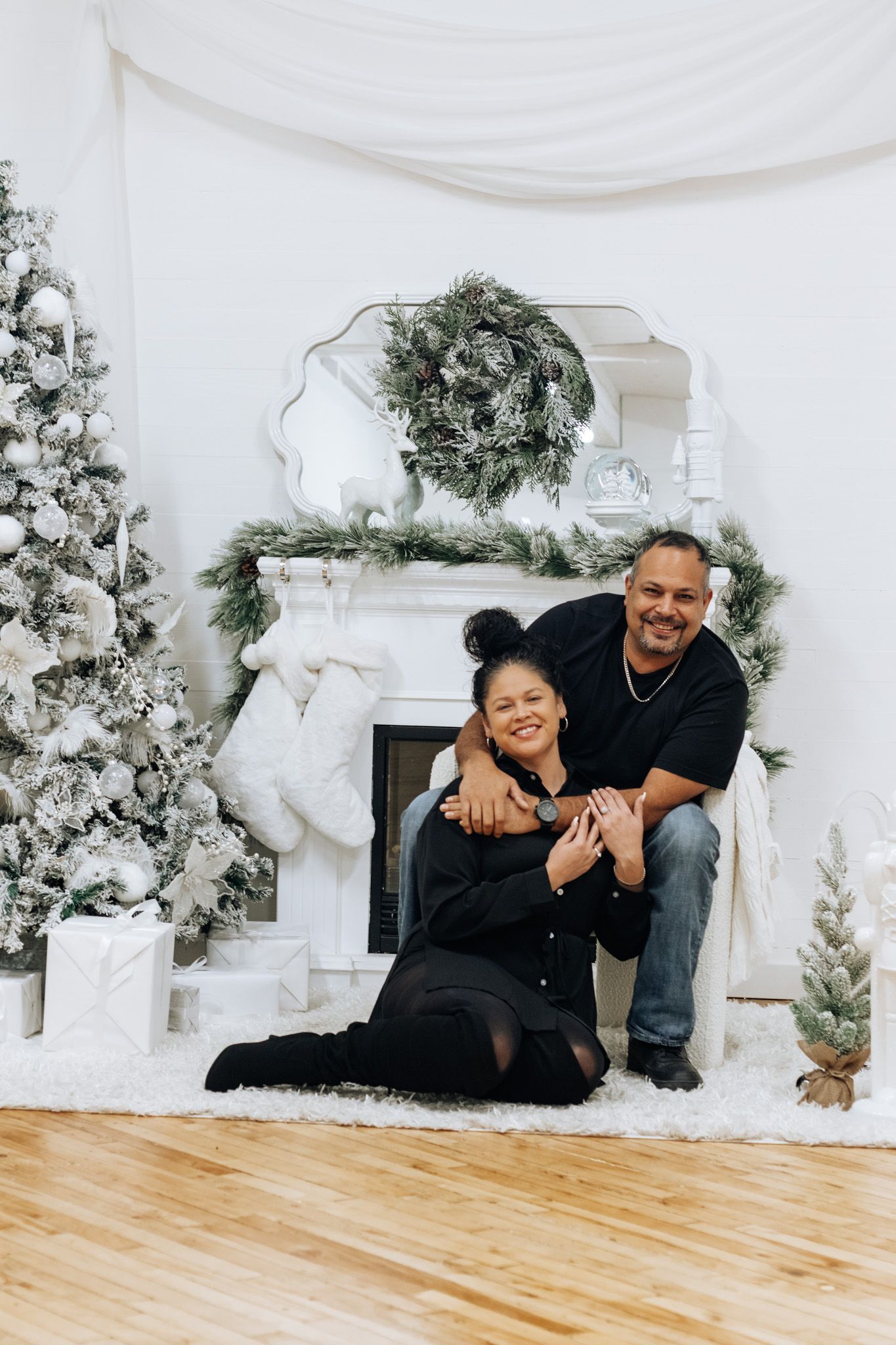 A couple shares a playful moment in front of a decorated white Christmas fireplace and flocked tree.