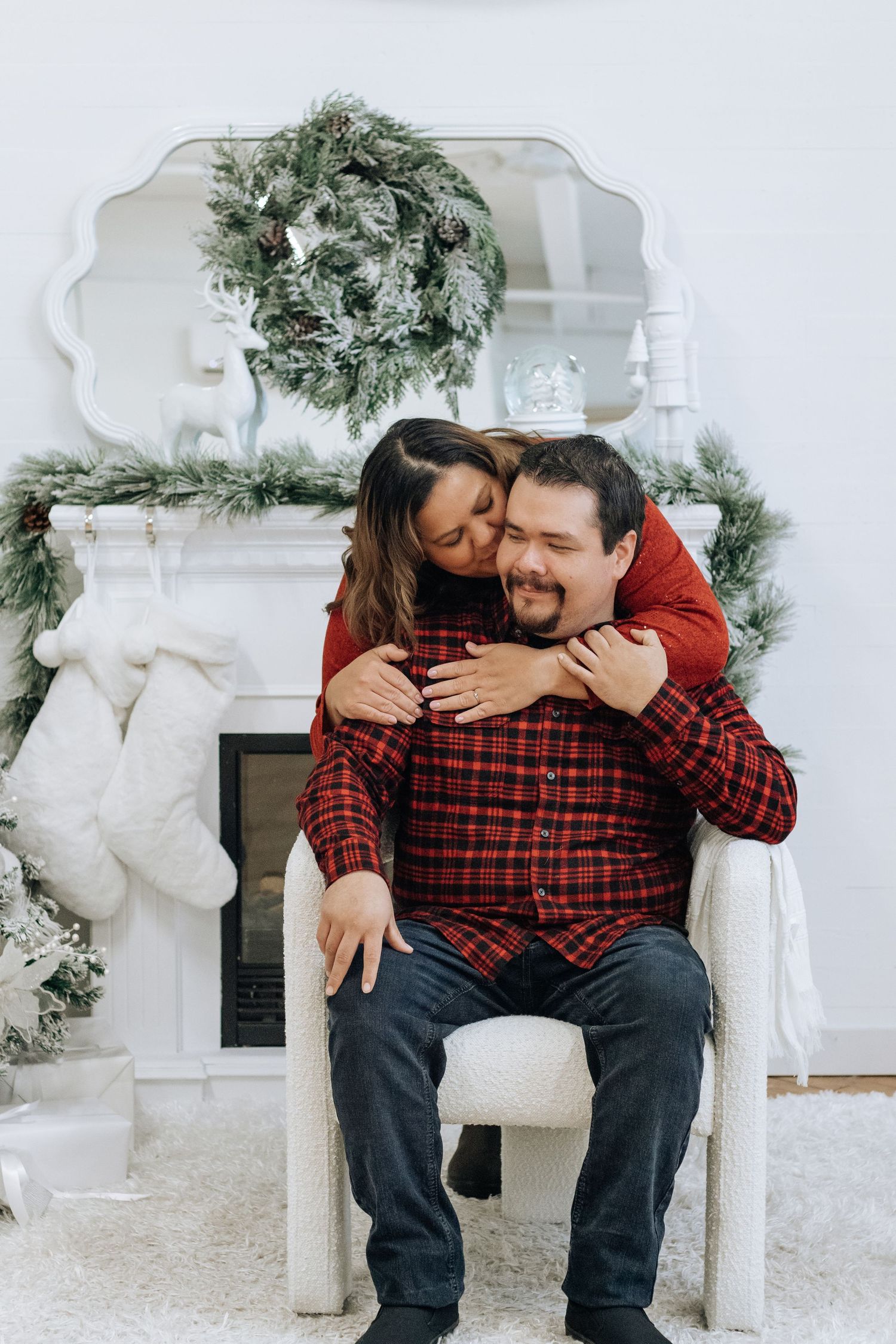 Couple embracing in chair by decorated Christmas fireplace with festive wreath.