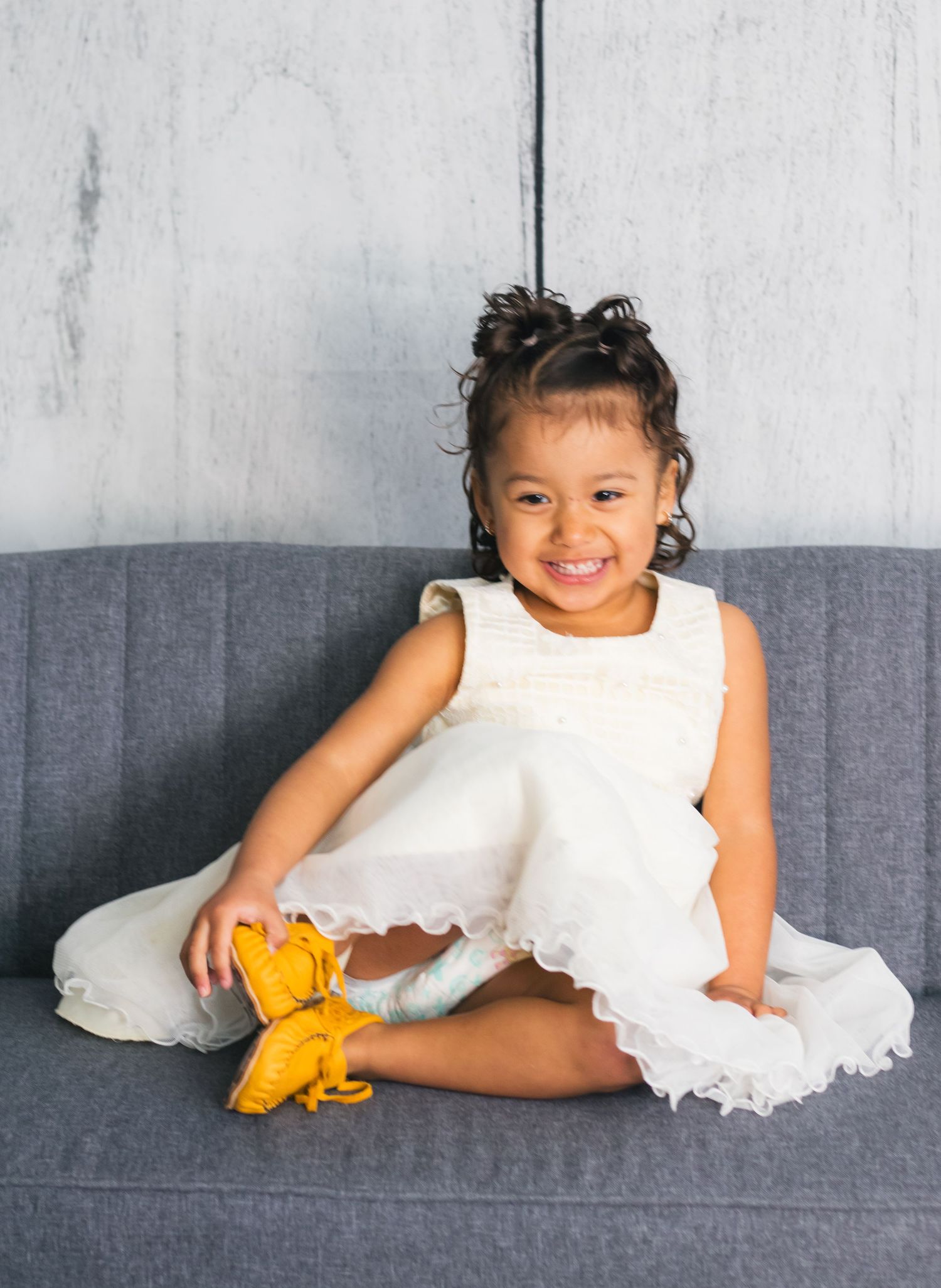 Child in white dress laying on grey couch playing with yellow dinosaur toy.