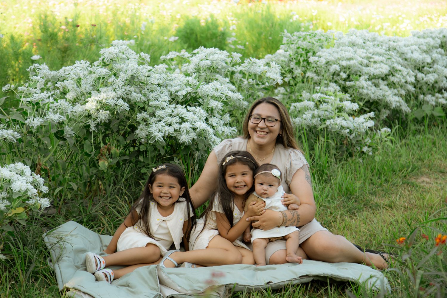 Family sitting together on blanket surrounded by white flowering bushes on sunny day.