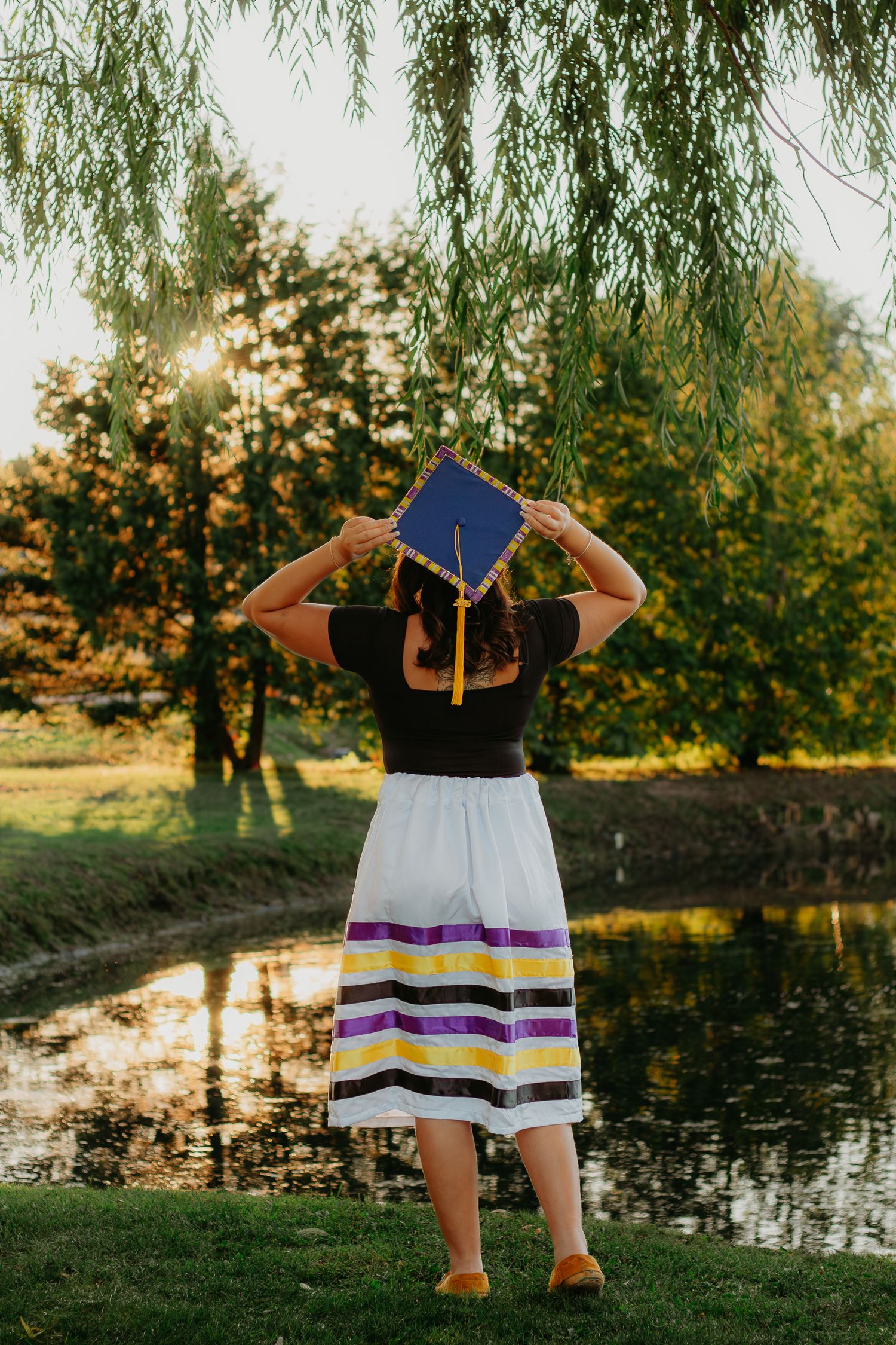 A graduate in cap and gown poses by a scenic pond with willow trees at sunset.