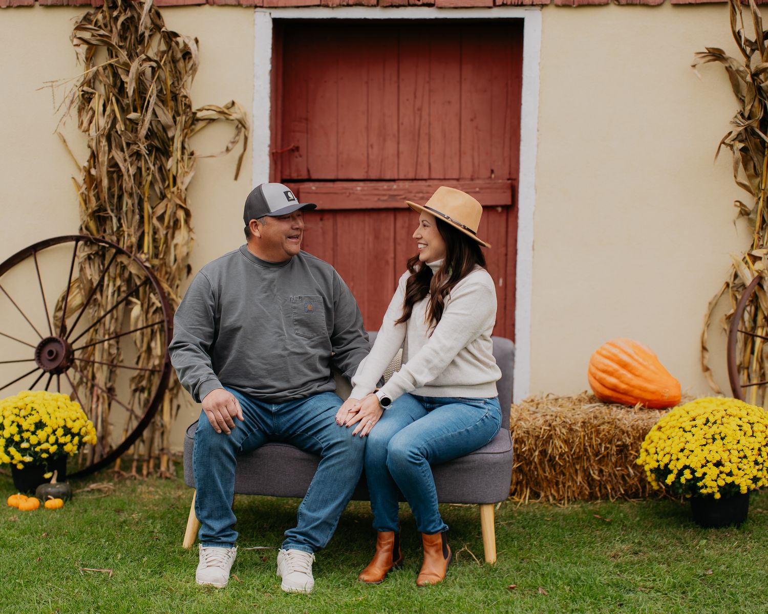 People relaxing on benches outside a barn decorated with cornstalks and yellow mums.
