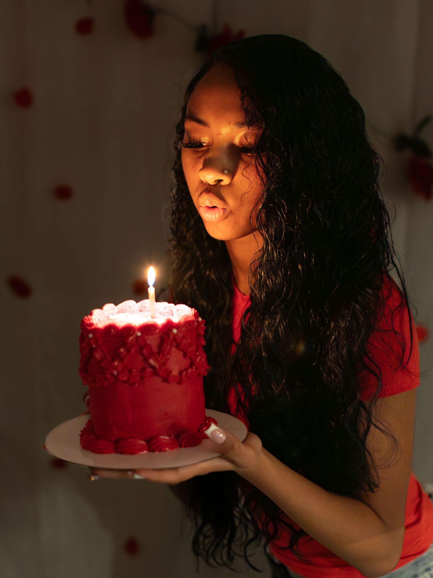 A red birthday cake with a lit candle being held in candlelight against a dark background.
