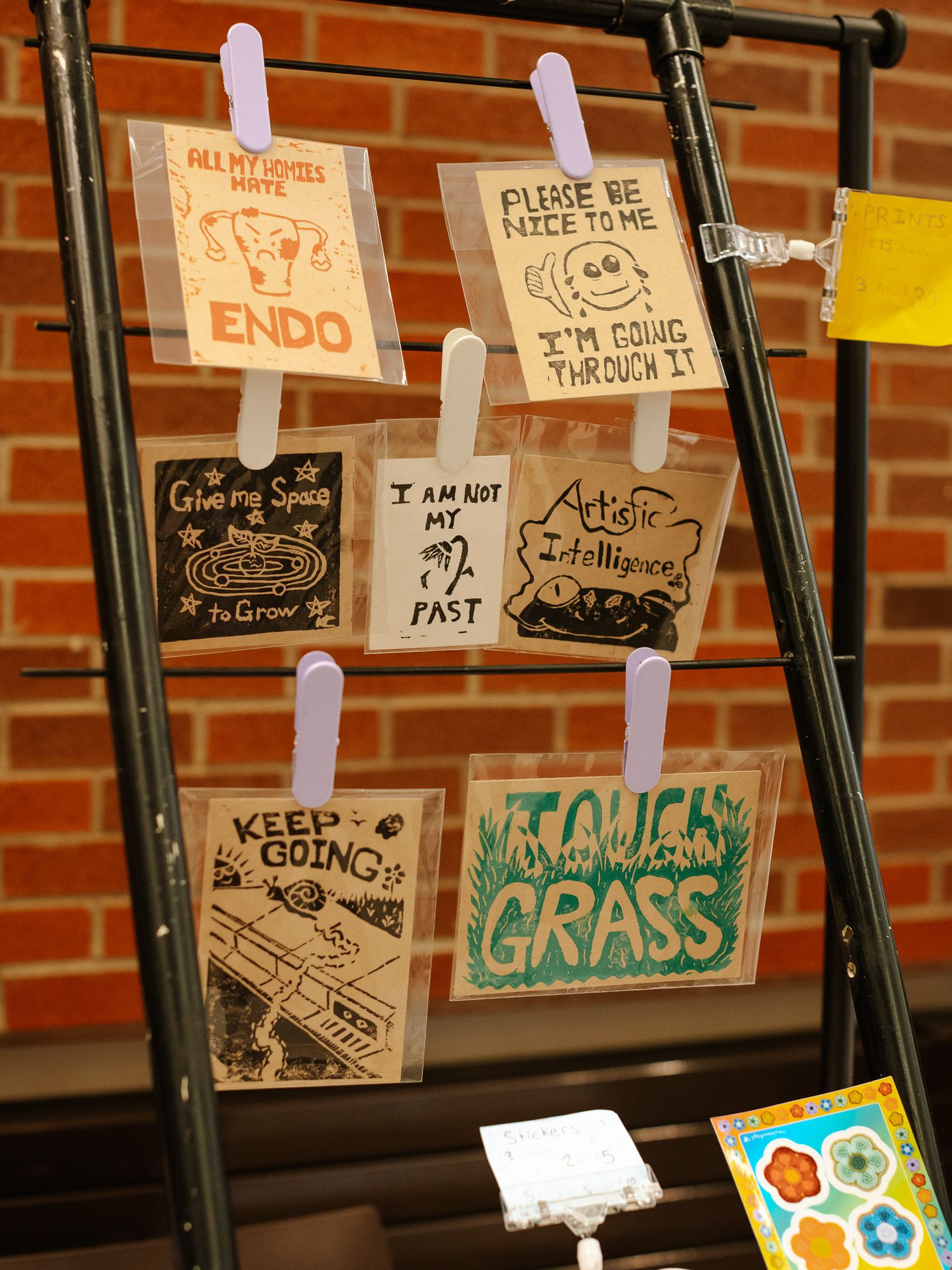Handmade motivational art cards with messages like Keep Going and Touch Grass displayed on a black rack with clothespins.