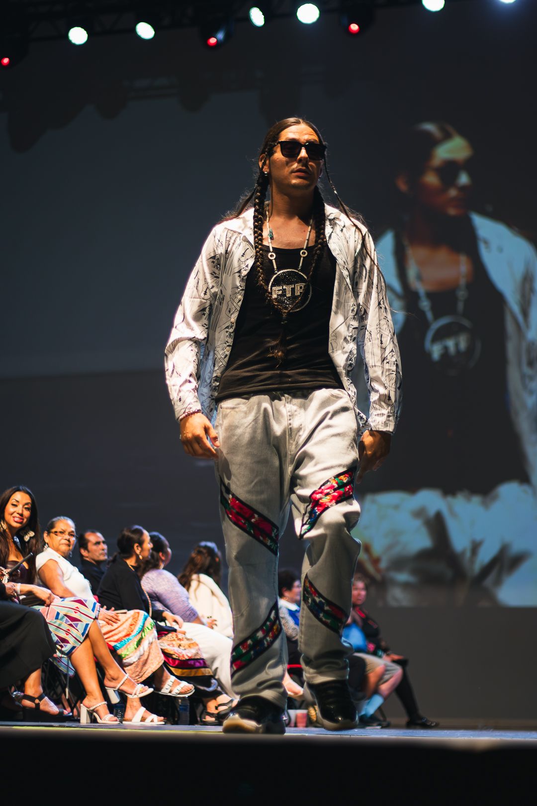 Model walks runway in silver jacket, FTF tee, and colorful-trimmed gray pants at a fashion show.