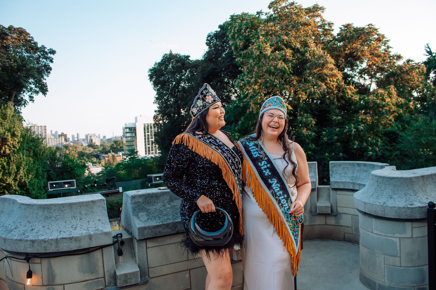Two women in Indigenous ceremonial regalia and sashes laugh together at outdoor evening event.