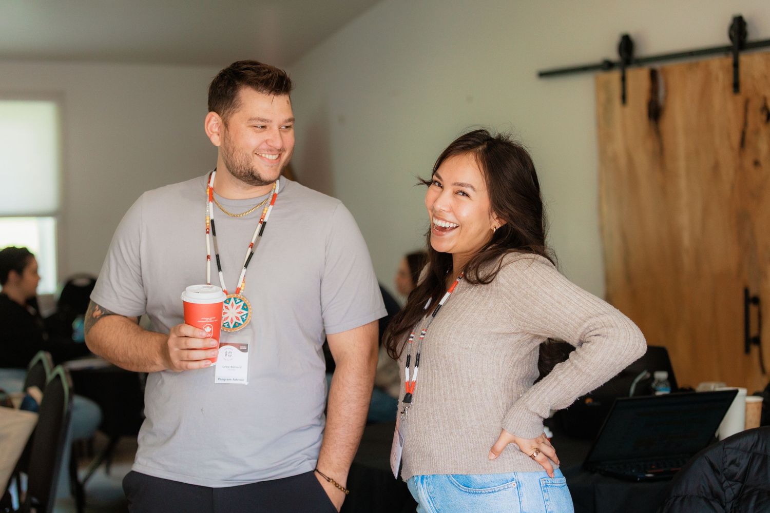 Two smiling colleagues wearing lanyards chat at an indoor networking event, one holding a festive drink.