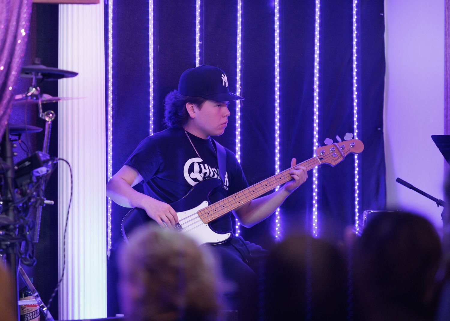 Musician playing electric bass guitar on stage with purple LED backdrop lighting at an indoor venue.