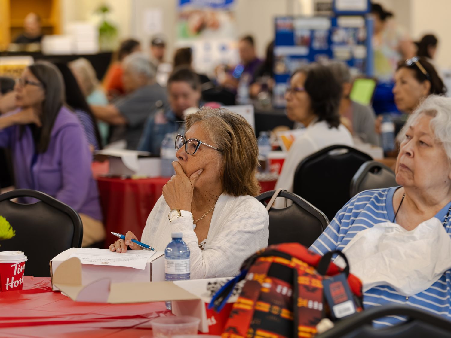 Indigenous women seated at tables listening attentively during a large community conference event.