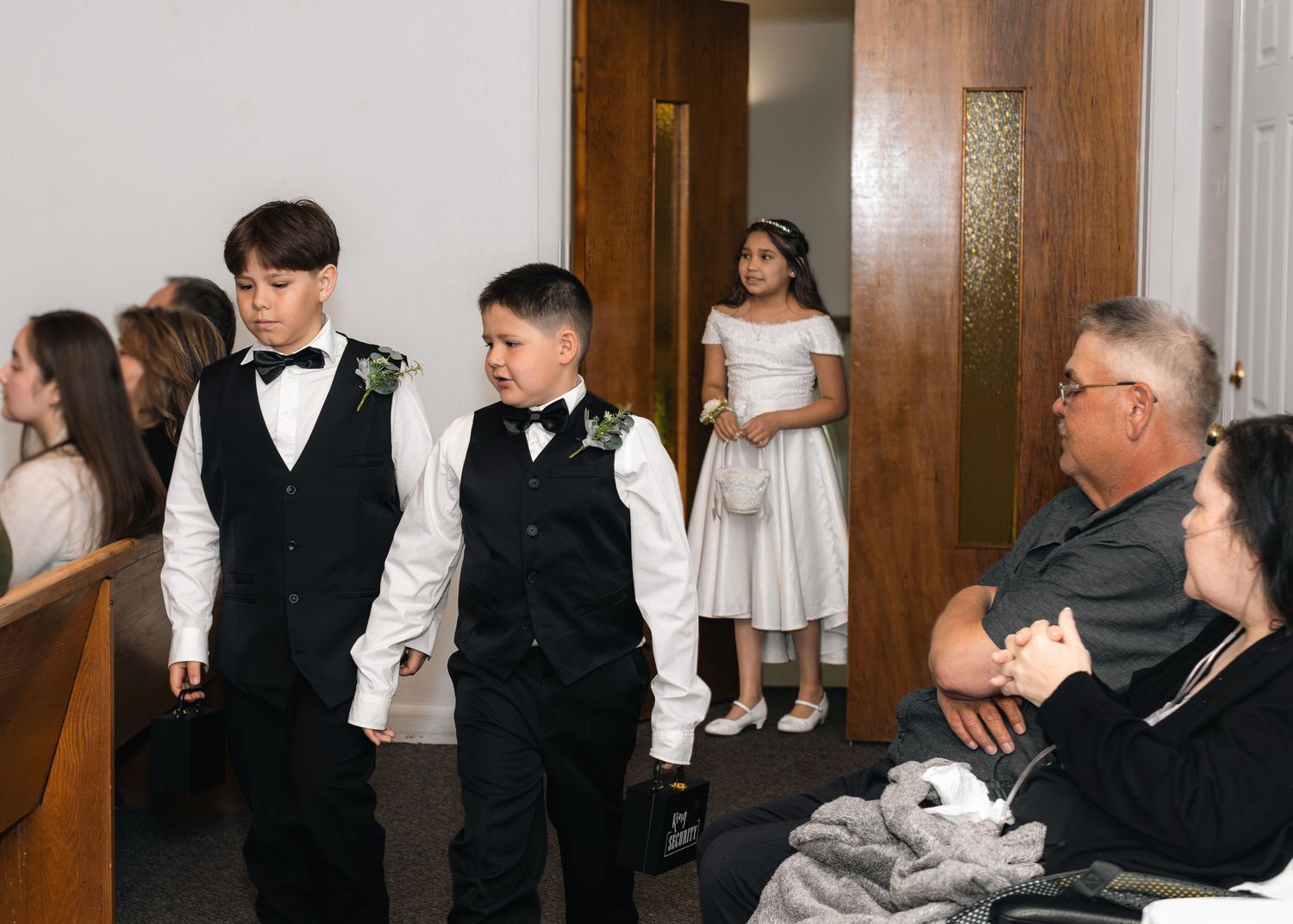 Young ring bearers in vests and bow ties walk down church aisle as flower girl and guests look on.