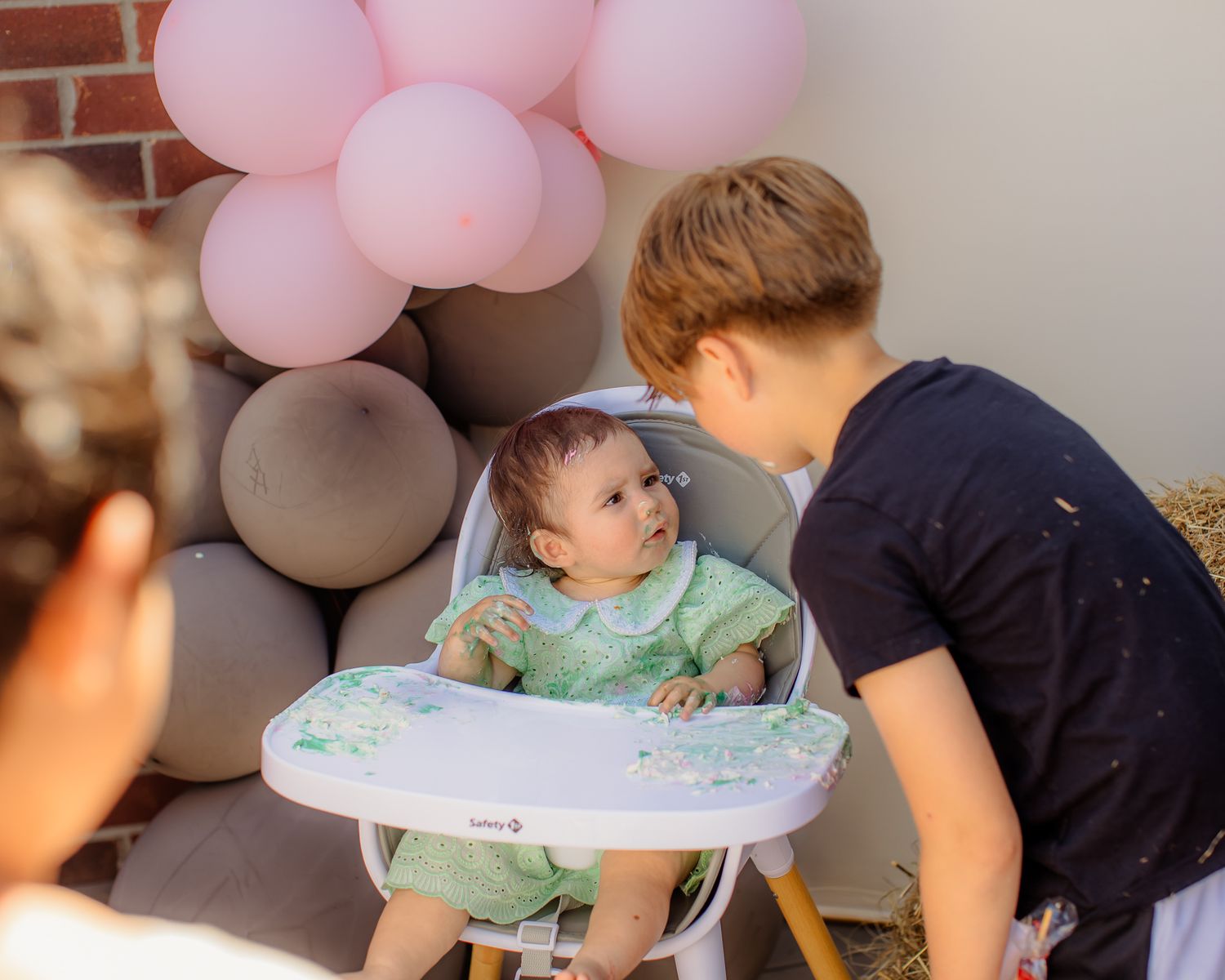 Young boy leaning in to interact with baby sitting in white high chair surrounded by pink and gray balloons.