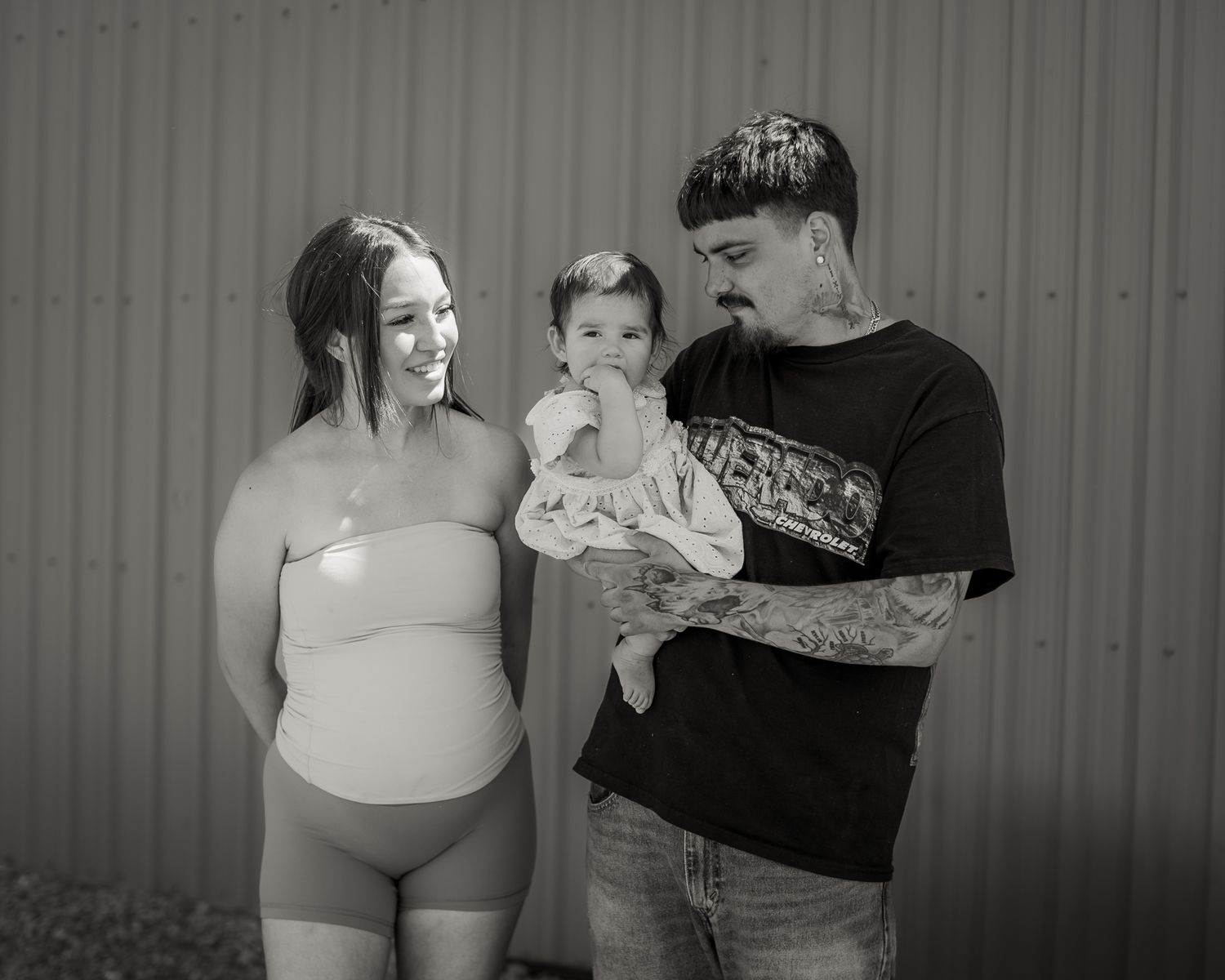 Black and white maternity photo of pregnant woman, tattooed man holding baby, family portrait outdoors.