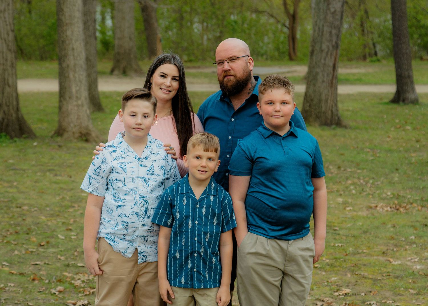 Family of five posing outdoors in a wooded park, parents with three boys wearing coordinating outfits in spring.