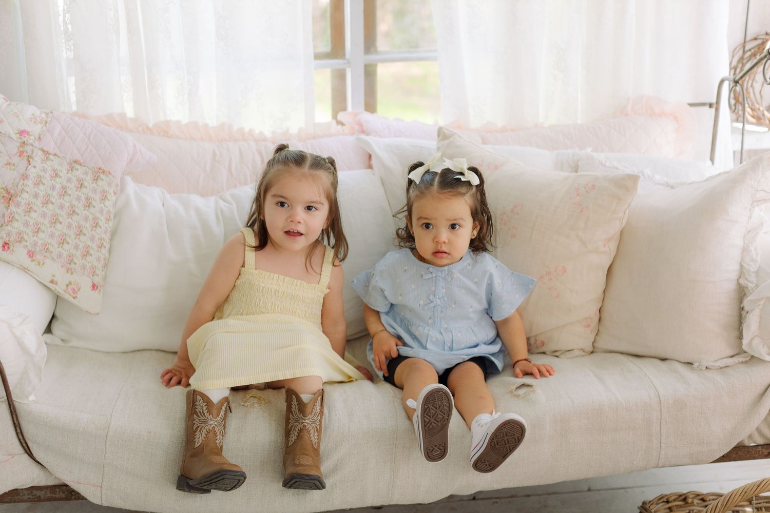 Two toddler girls in pastel dresses sitting on a white couch with floral pillows in a bright, airy room.