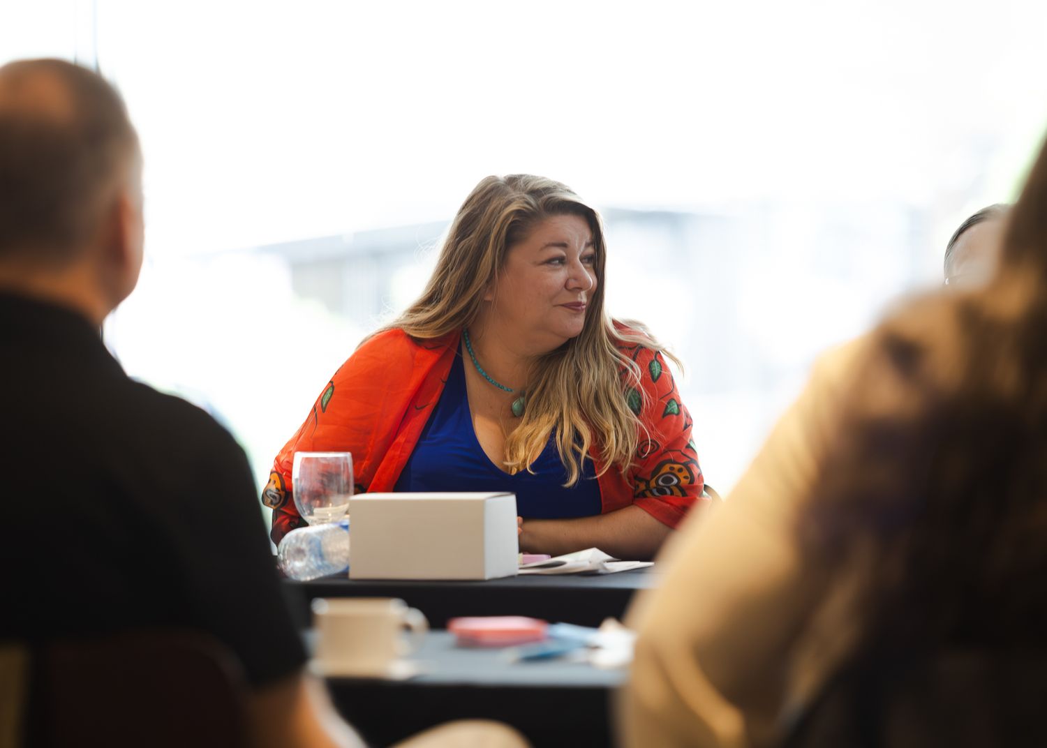 Woman in red blazer and blue top sits at a conference table, engaged in a meeting discussion.