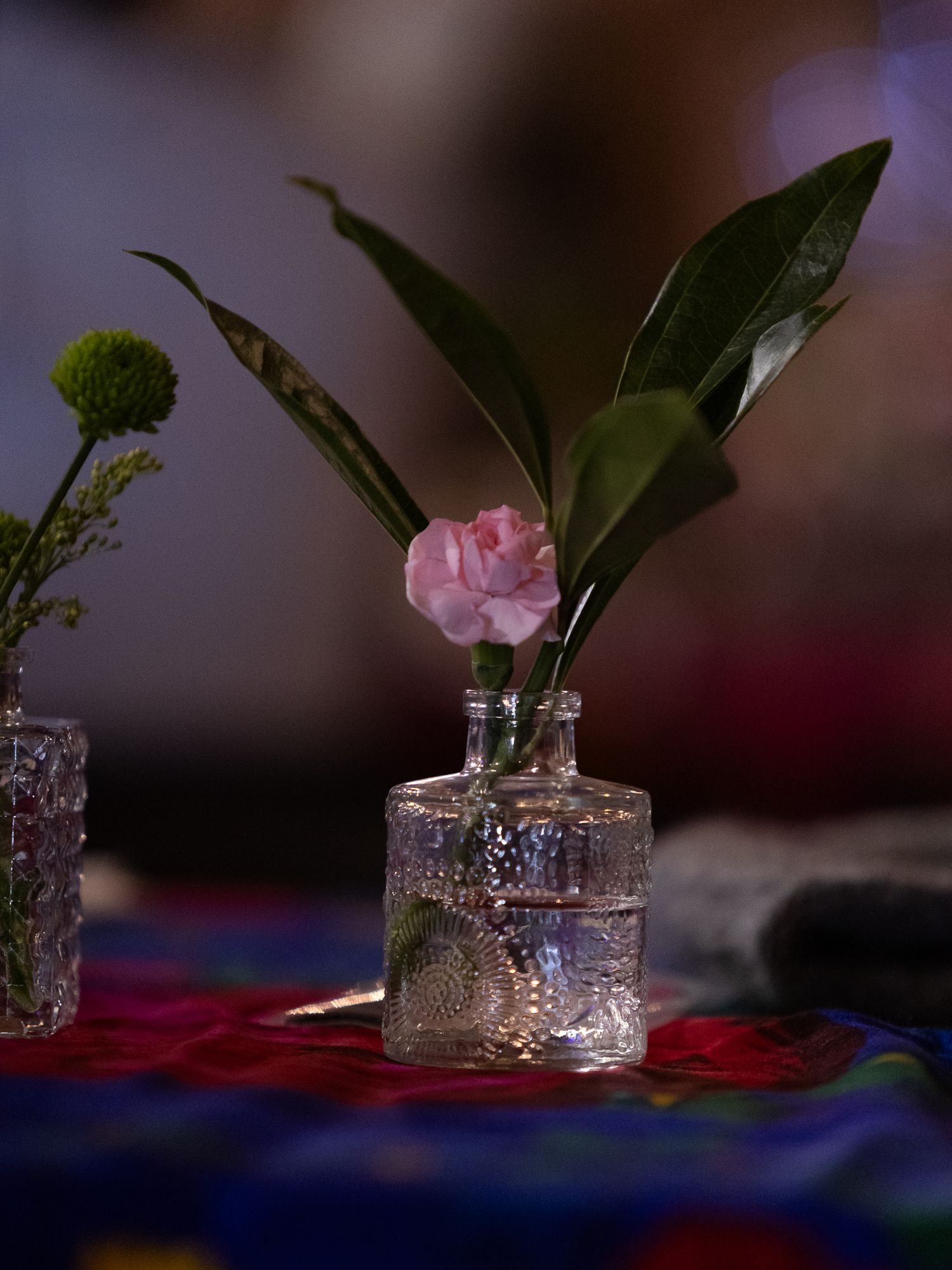 Pink carnation and green leaves in a clear glass bottle vase on a colorful tablecloth.