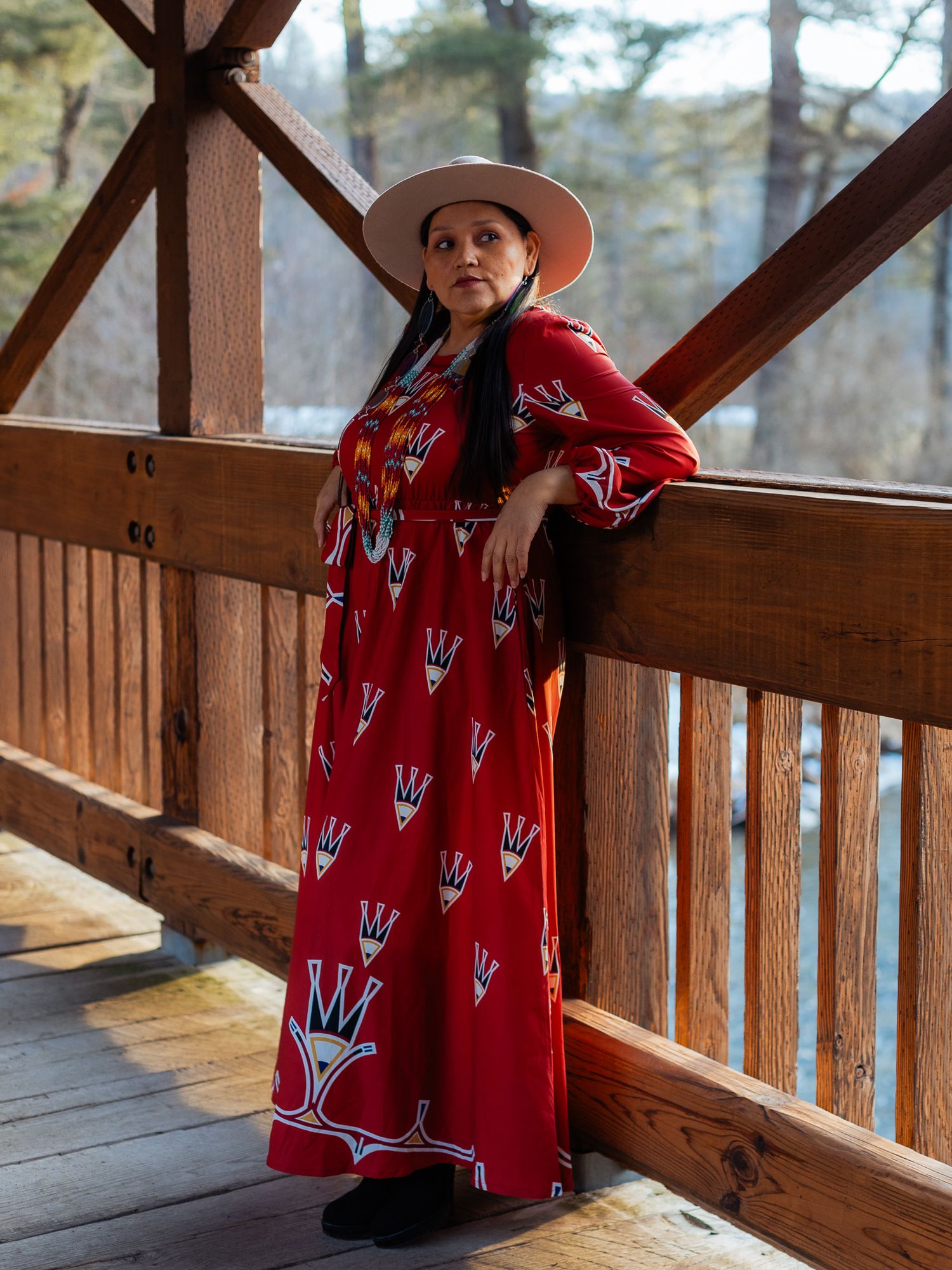 Woman in red crown-print maxi dress and tan wide-brim hat poses on wooden deck surrounded by trees.