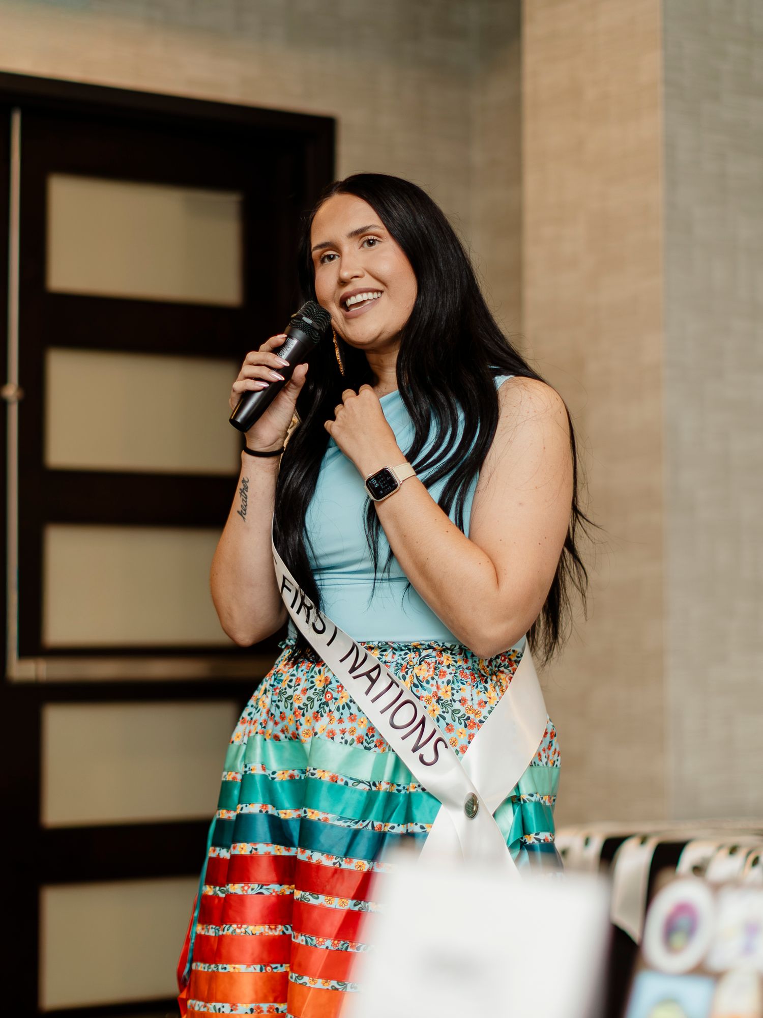 Woman wearing a First Nations sash speaks into a microphone in a colorful skirt and teal top.