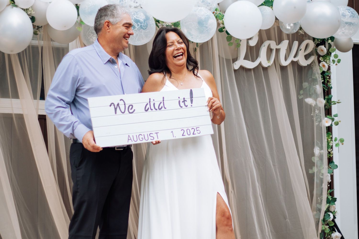 Newlyweds laugh holding a We Did It sign dated August 1 2025 at decorated outdoor wedding venue.