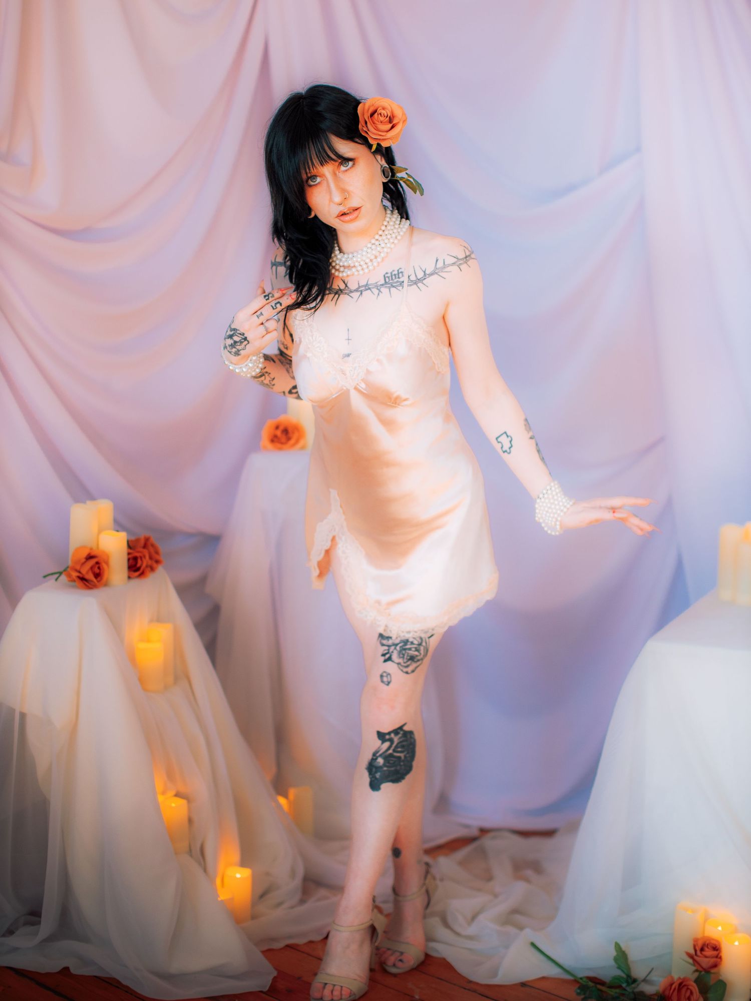 Woman in silk slip dress with tattoos poses among candles and orange roses against draped white fabric backdrop.