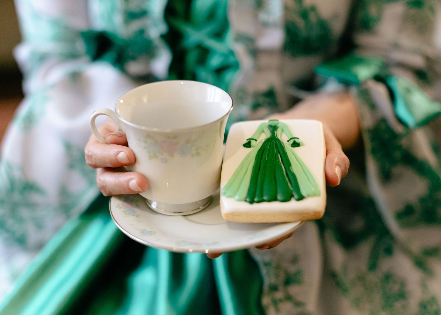Person holding a fine china teacup and saucer with a decorated green wedding dress sugar cookie.
