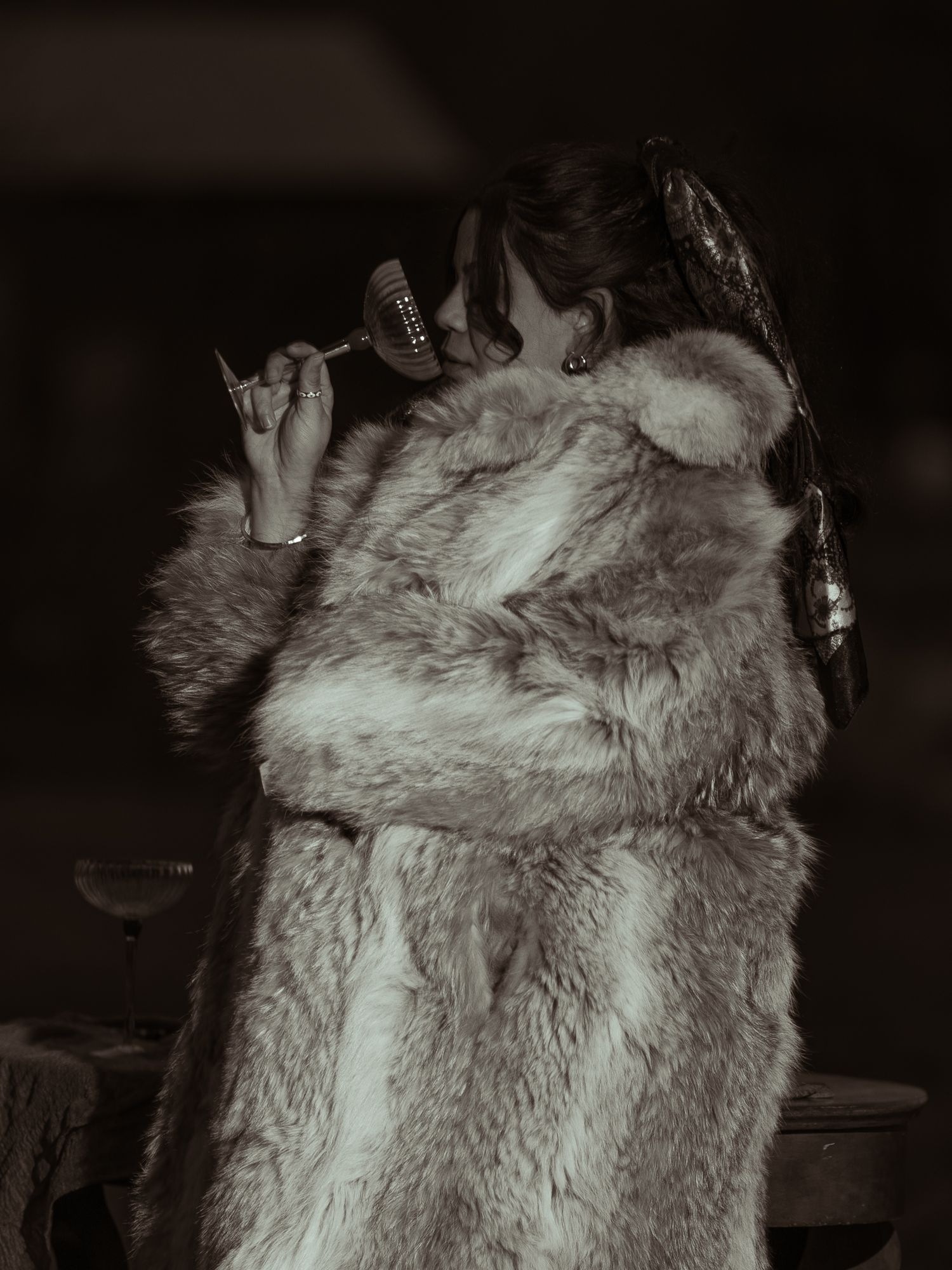 Black and white photo of woman in fur coat drinking from a wine glass outdoors.