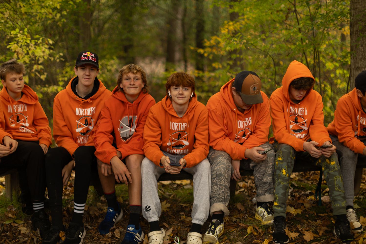 Line of people in bright orange sweatshirts sitting on fallen logs in autumn forest.