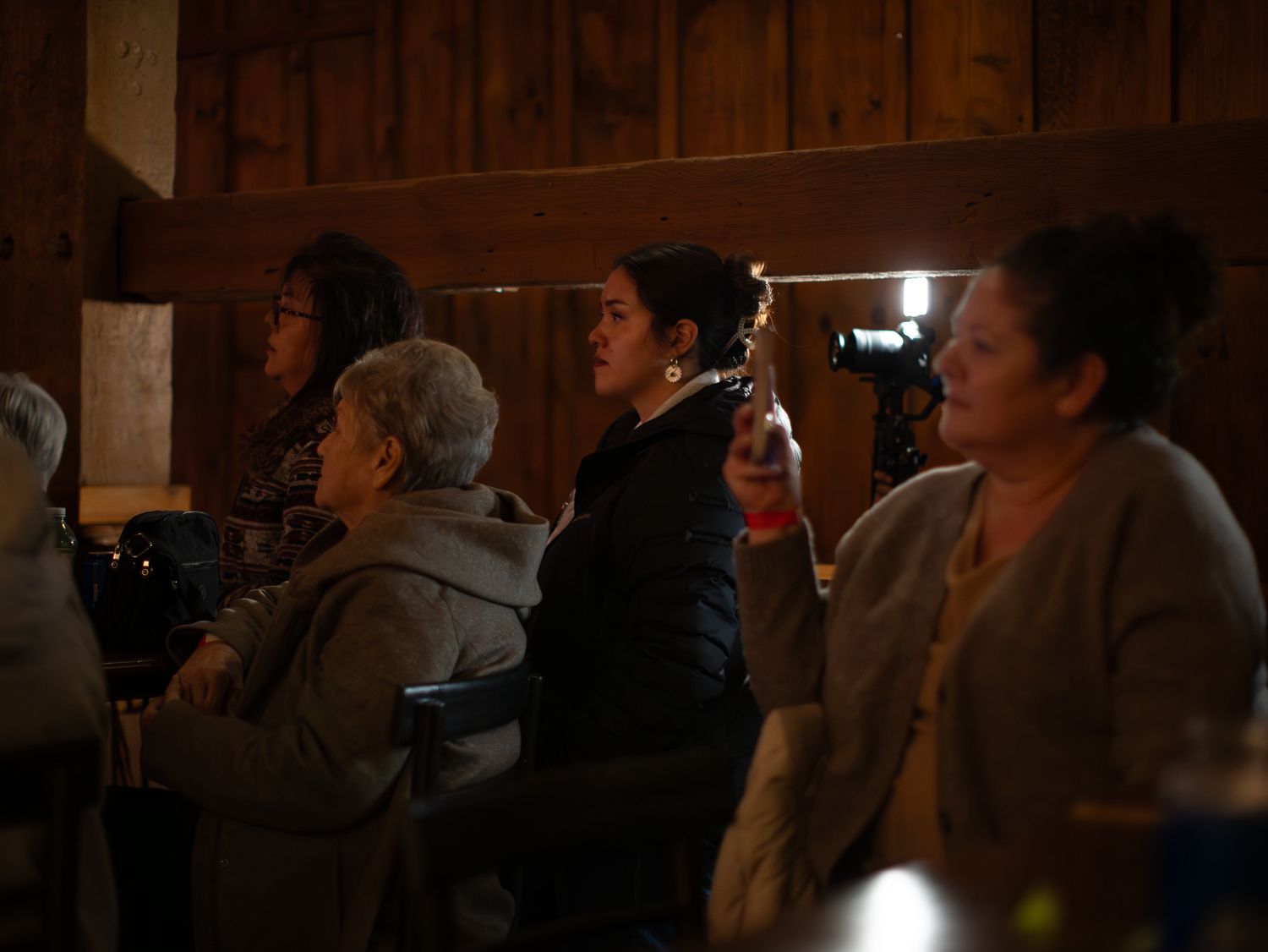 Audience members watching a performance in a rustic venue with wooden walls and dim lighting.