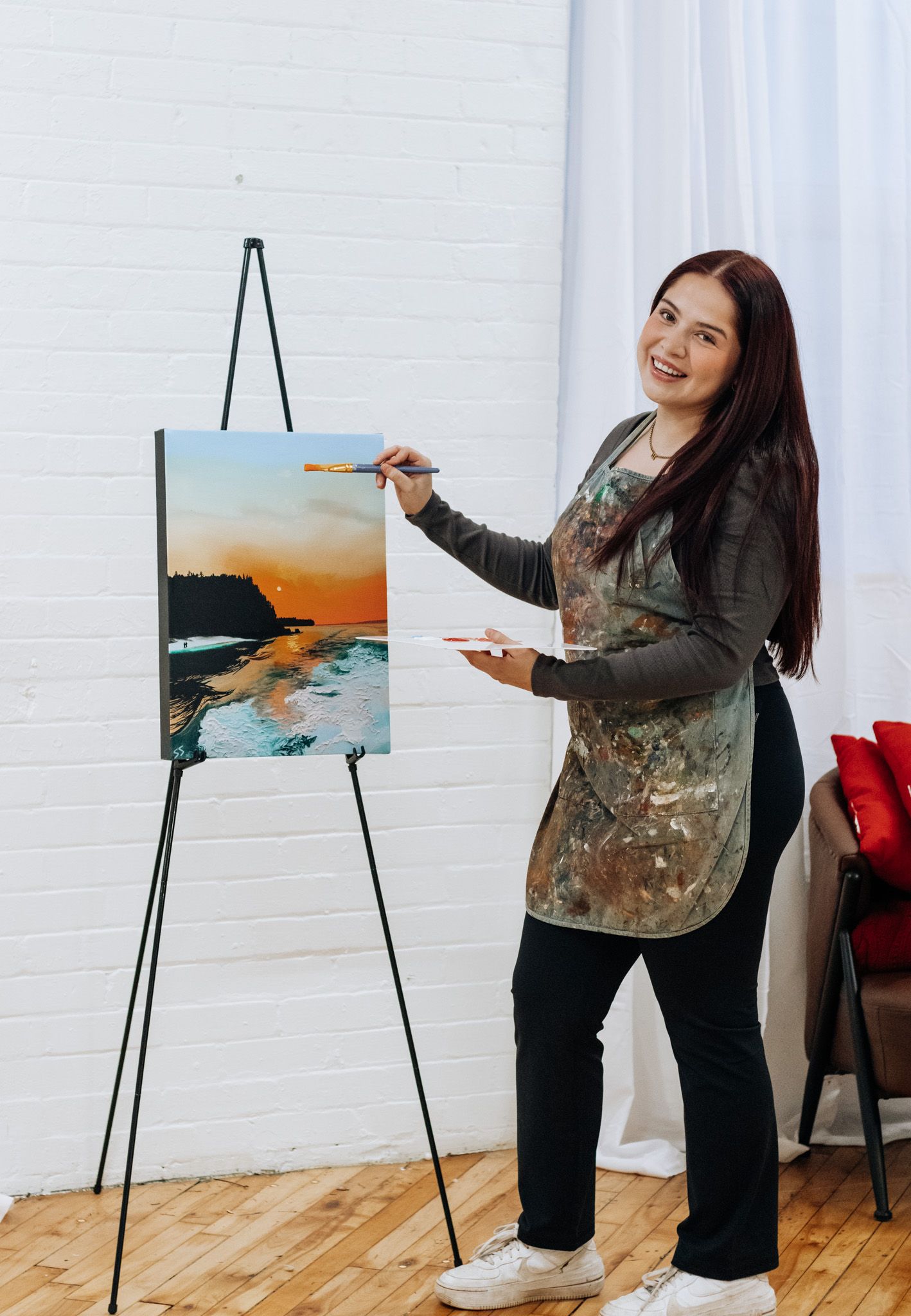 Artist in paint-splattered smock works on landscape painting at easel in bright studio space.