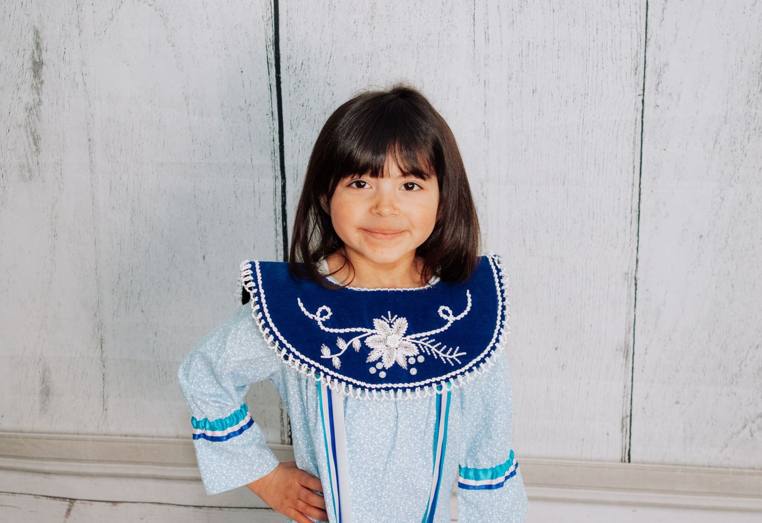 A young child wearing a blue and white traditional dress with embroidered details poses against a light background.