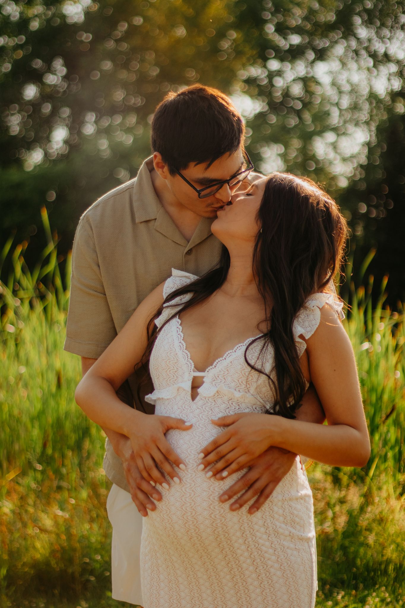 A romantic maternity photoshoot in a sunlit field with a couple embracing in white and beige outfits.