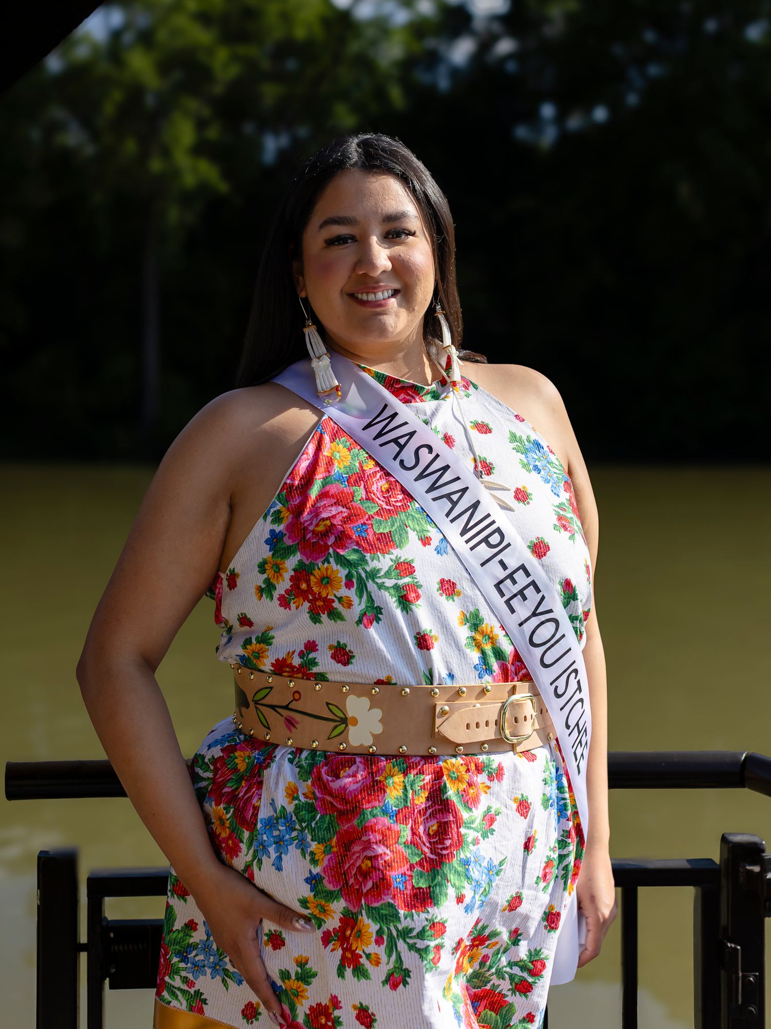 Young woman wearing a floral jumpsuit and pageant sash smiles outdoors near a scenic waterway.