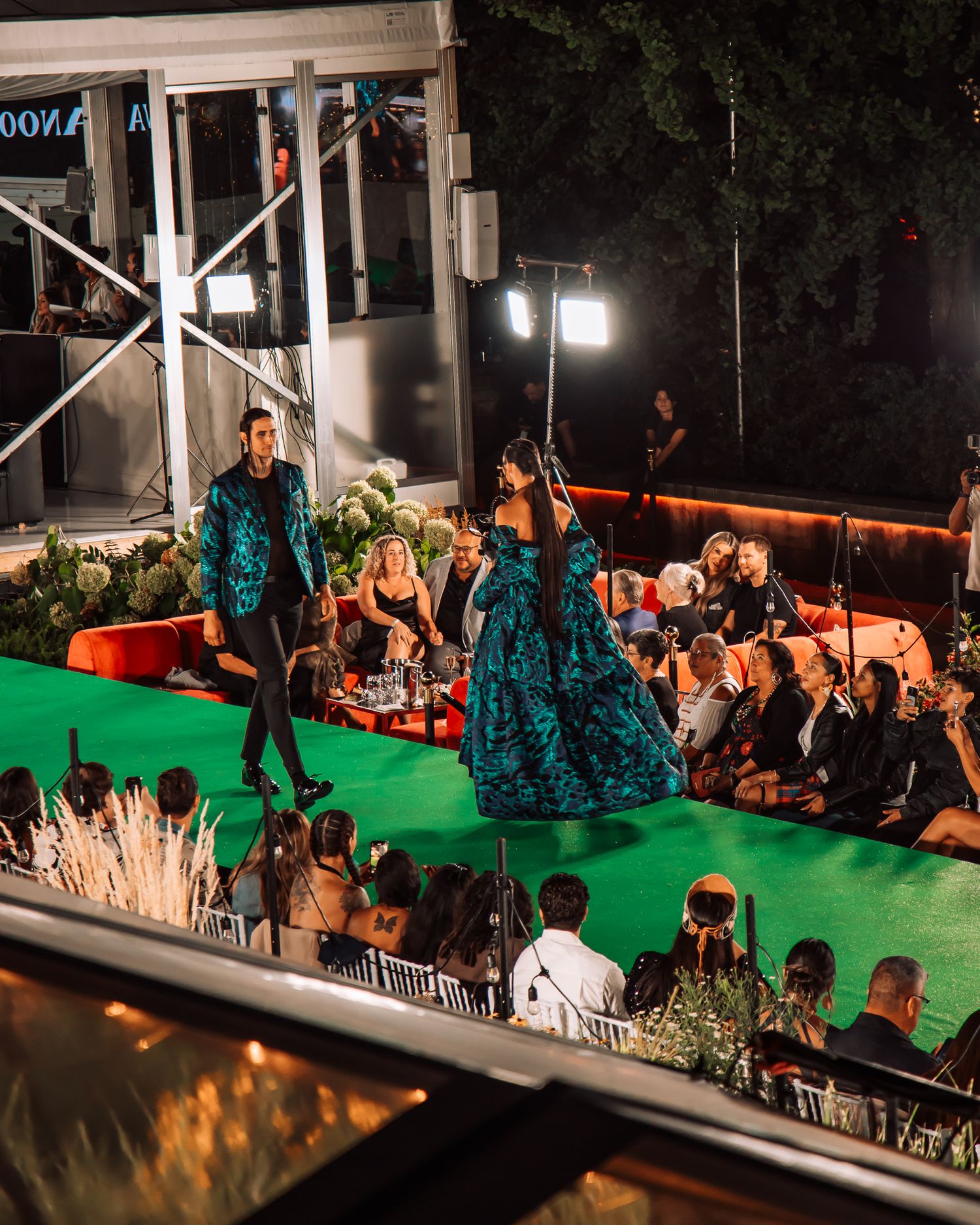 Two models in teal floral outfits walk green runway at an elegant outdoor night fashion show.
