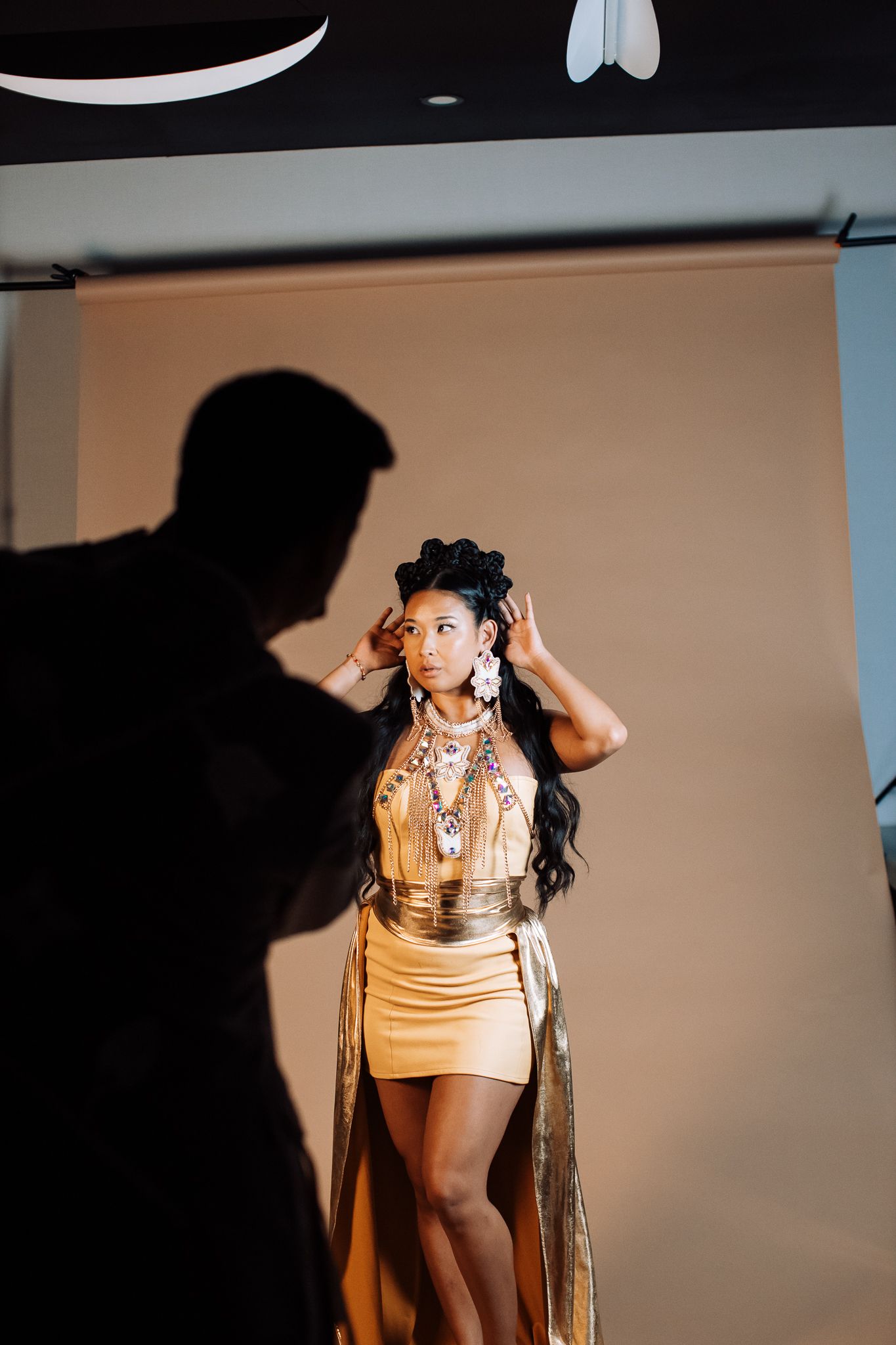 Model in gold mini dress poses during a professional photo shoot with photographer silhouette in foreground.