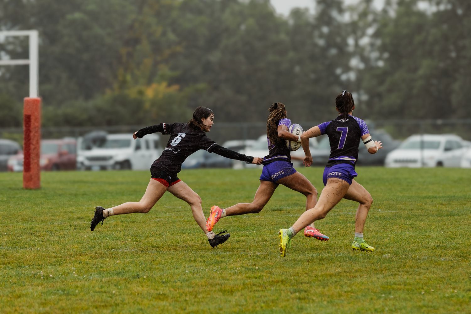 Women rugby players compete in a rainy outdoor match, with one player reaching to tag another during a fast-paced game.