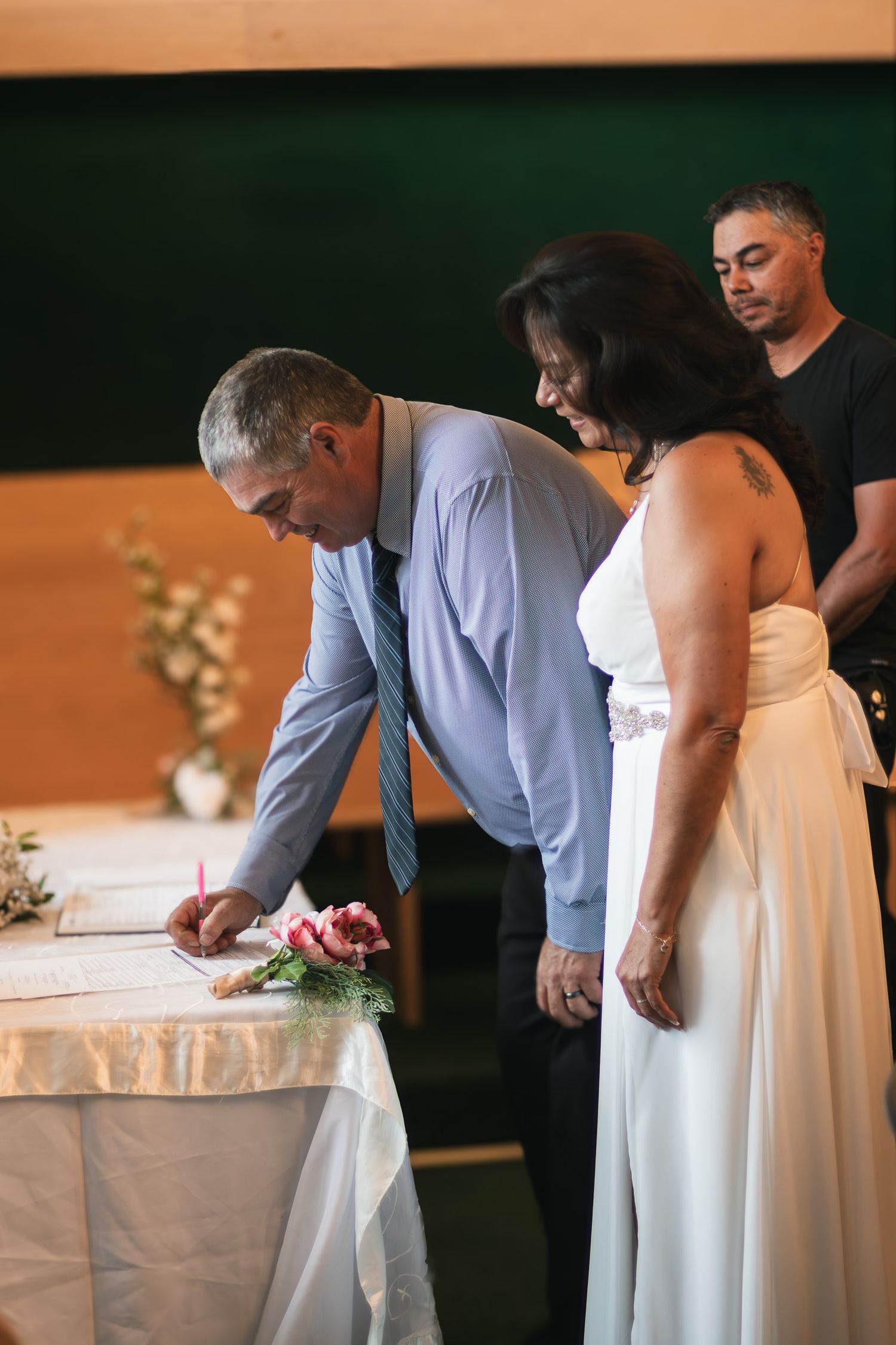 Bride and groom sign wedding register at ceremony table with witness standing behind them.