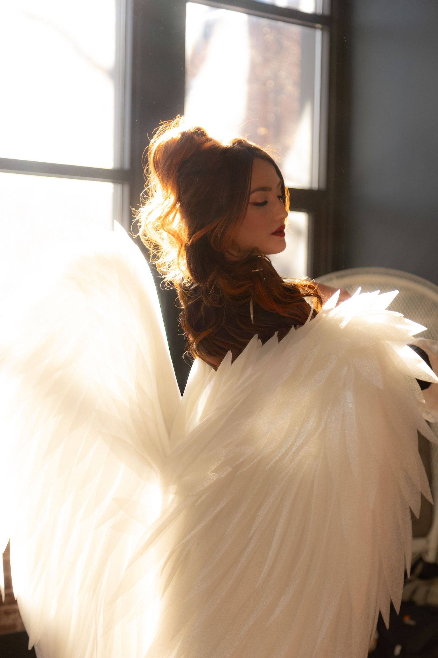 Young woman with angel wings posing in golden backlight near window, ethereal beauty photography.