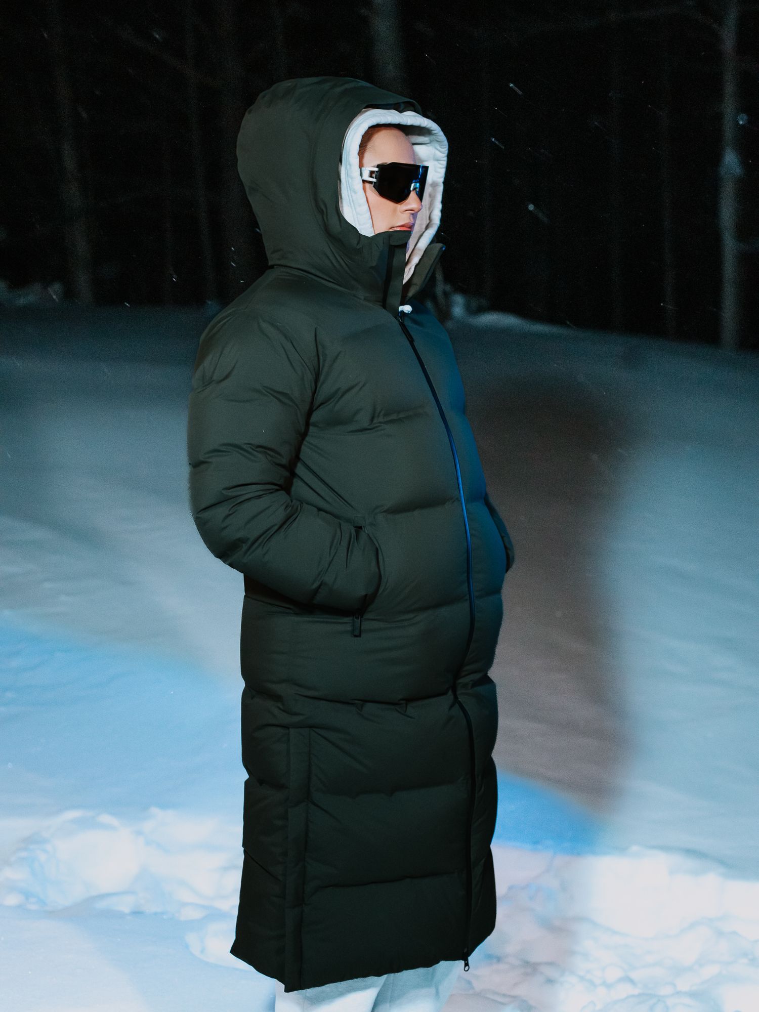 Person wearing a long dark green hooded puffer coat with sunglasses standing in snowy night setting.