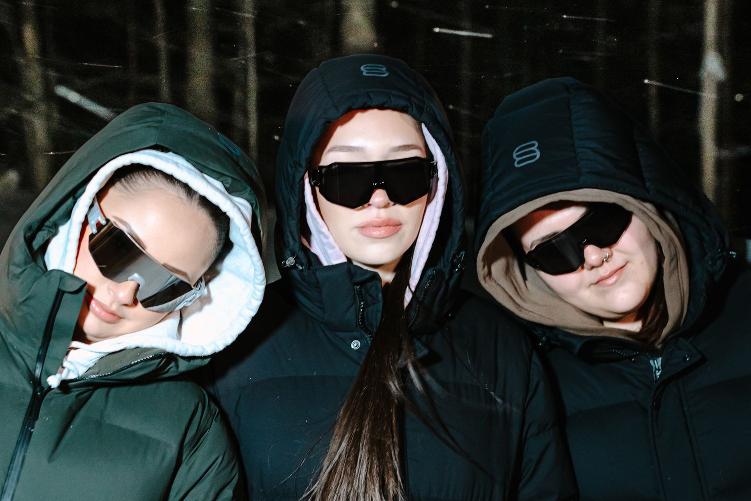 Three women in hooded puffer jackets and black sunglasses posing outdoors in a snowy winter setting.
