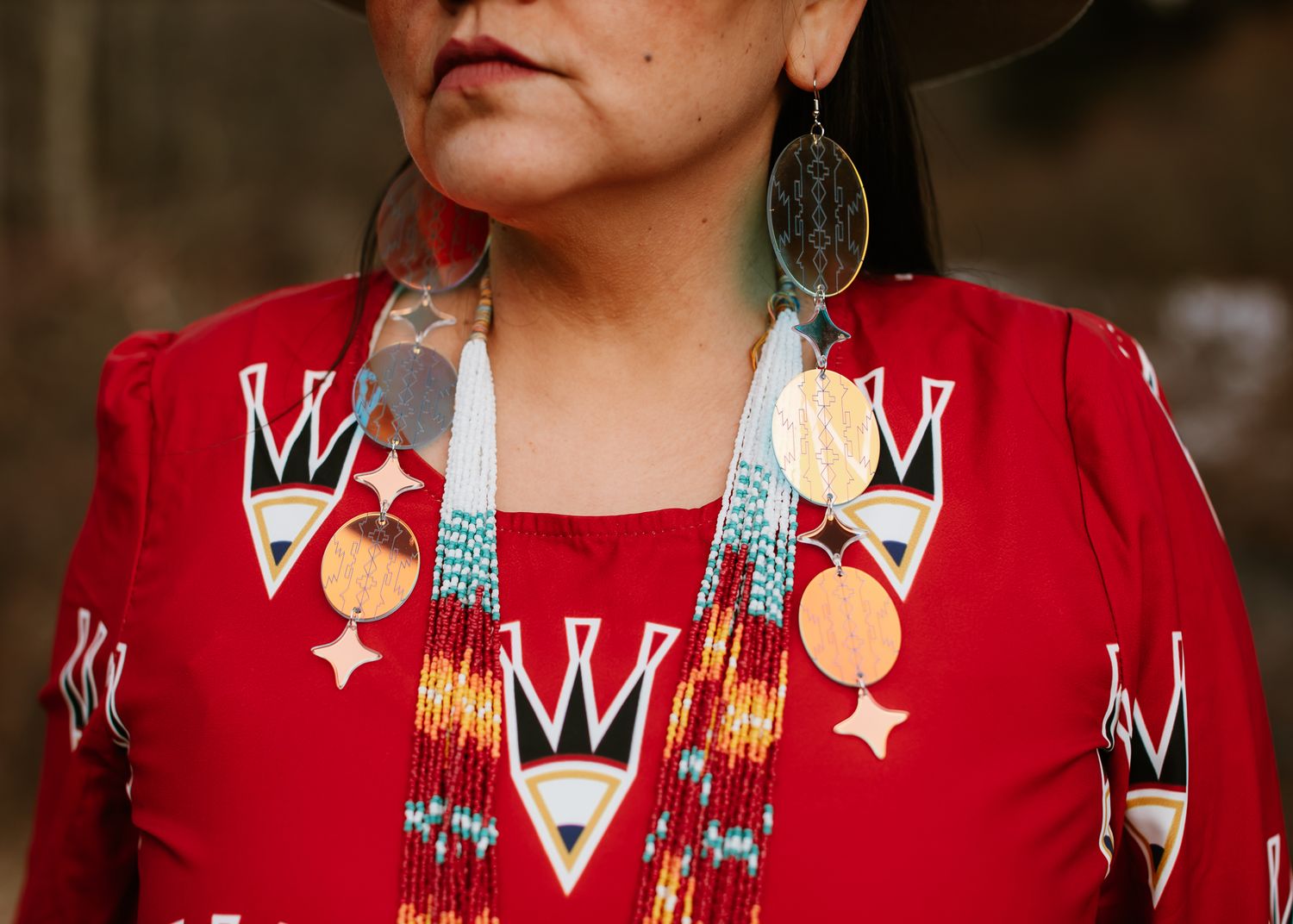 Native American woman in red traditional dress with beaded necklace and ornate earrings at outdoor gathering.