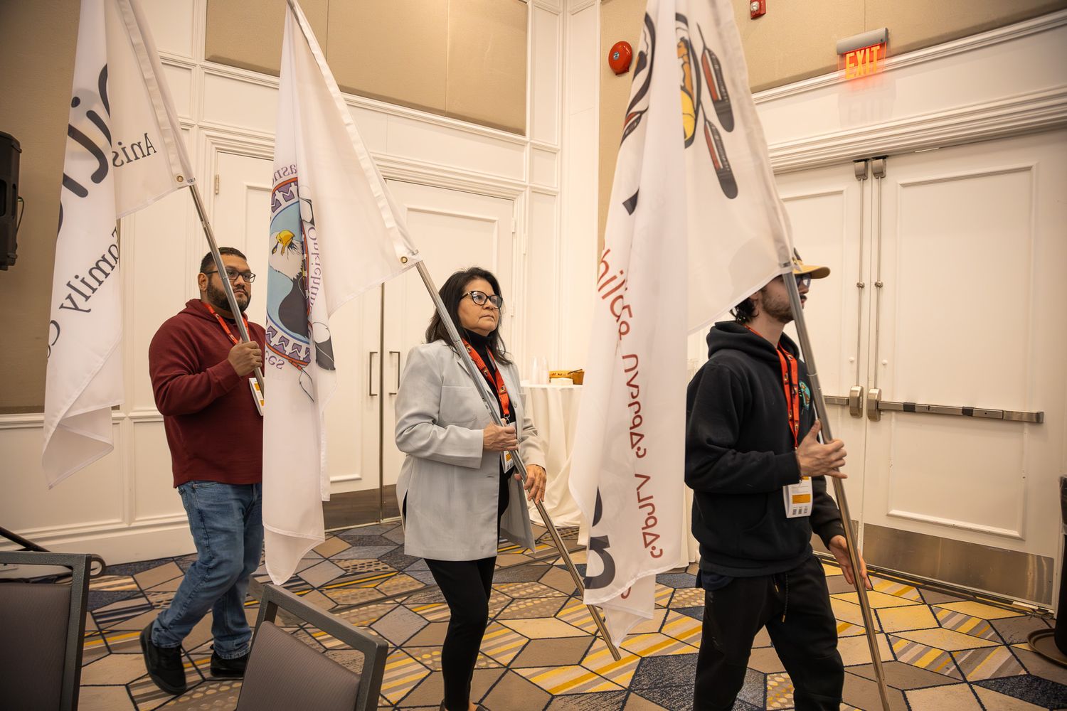 Three people carry ceremonial flags with Indigenous designs through a carpeted event hall.